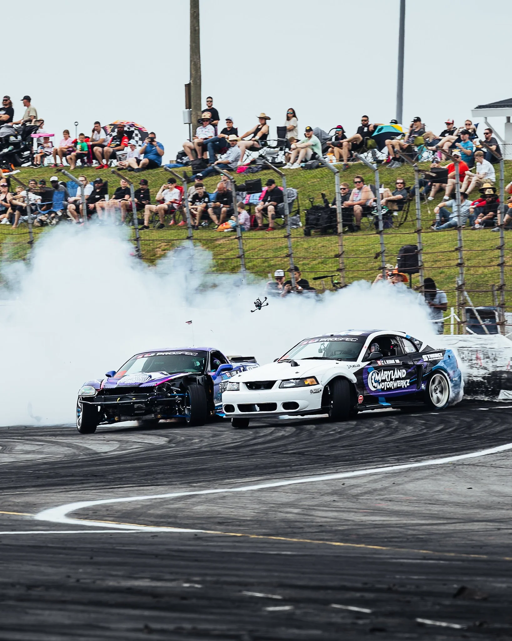 Two race cars are drifting on a track, creating smoke, with a crowd of spectators watching from a hill in the background.