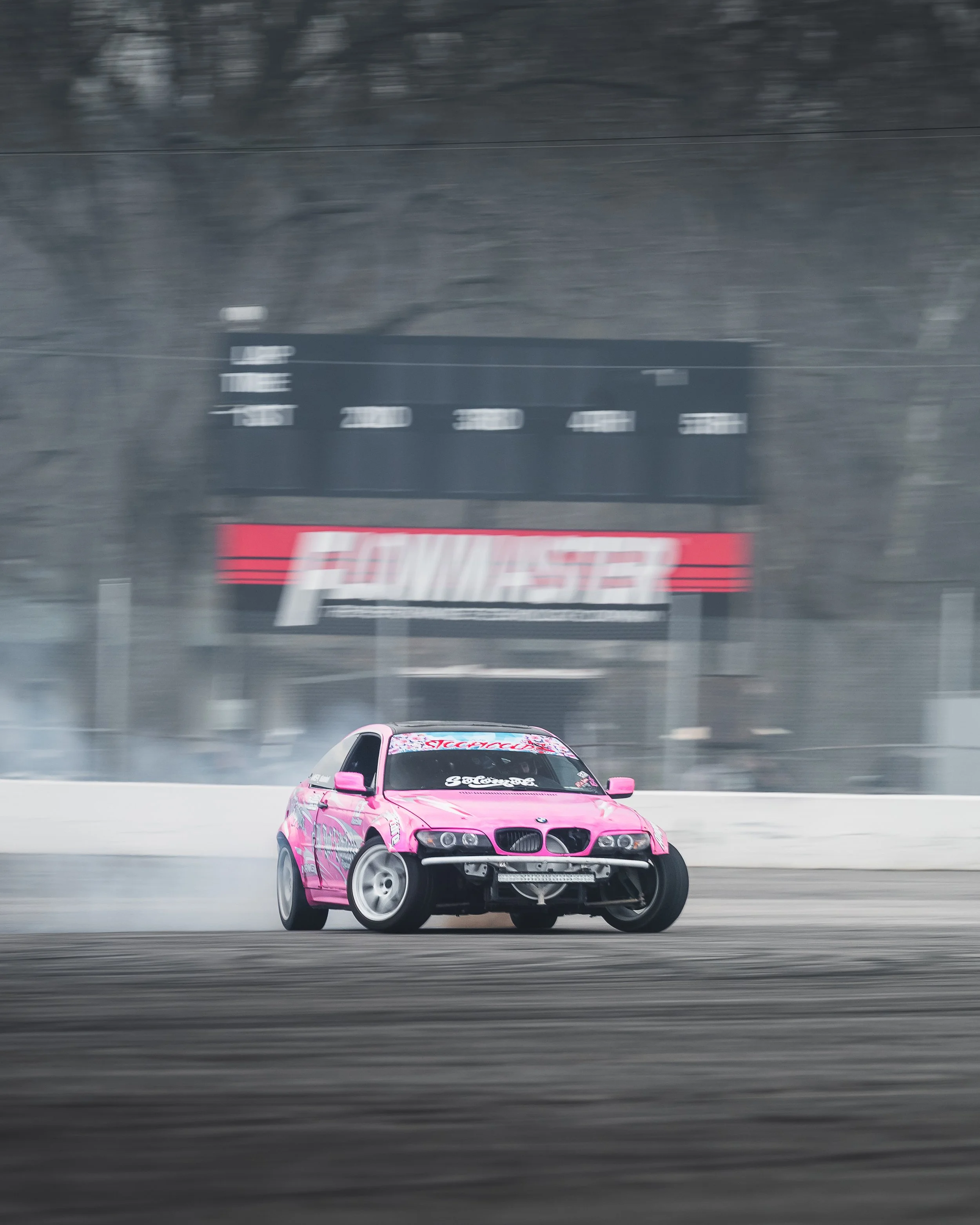 Pink race car drifting on a race track with tire smoke, blurred background of trees, and a sign that says 'Ford Mustang' in the distance.