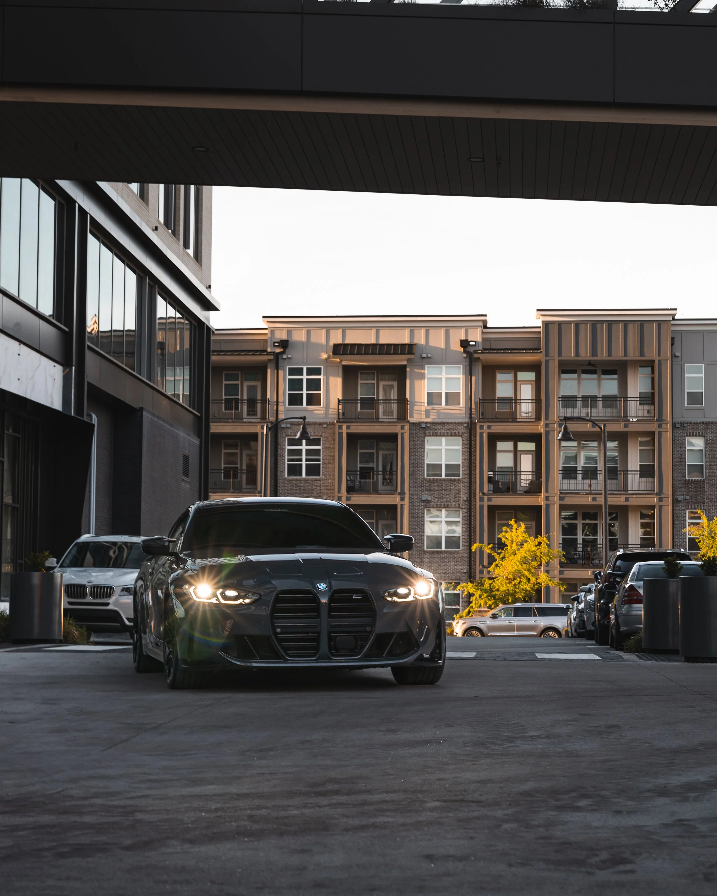 Black BMW car with headlights on parked in an urban parking lot, with modern apartment buildings and trees in the background.