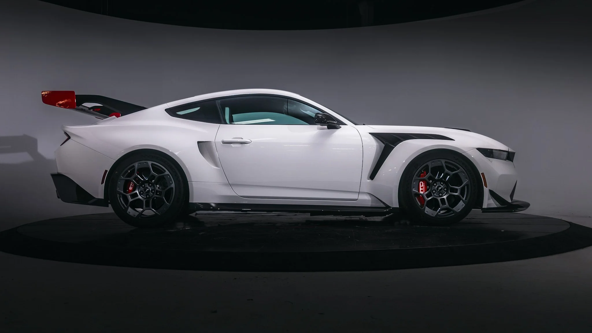 A white sports car with black and red accents, parked on a black circular platform against a plain background.