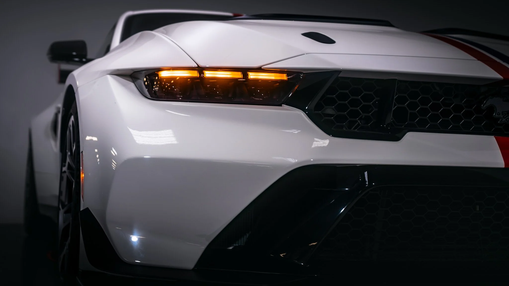 Front view of a white sports car with a red stripe, featuring a black honeycomb grille, sharp headlights, and aerodynamic design.