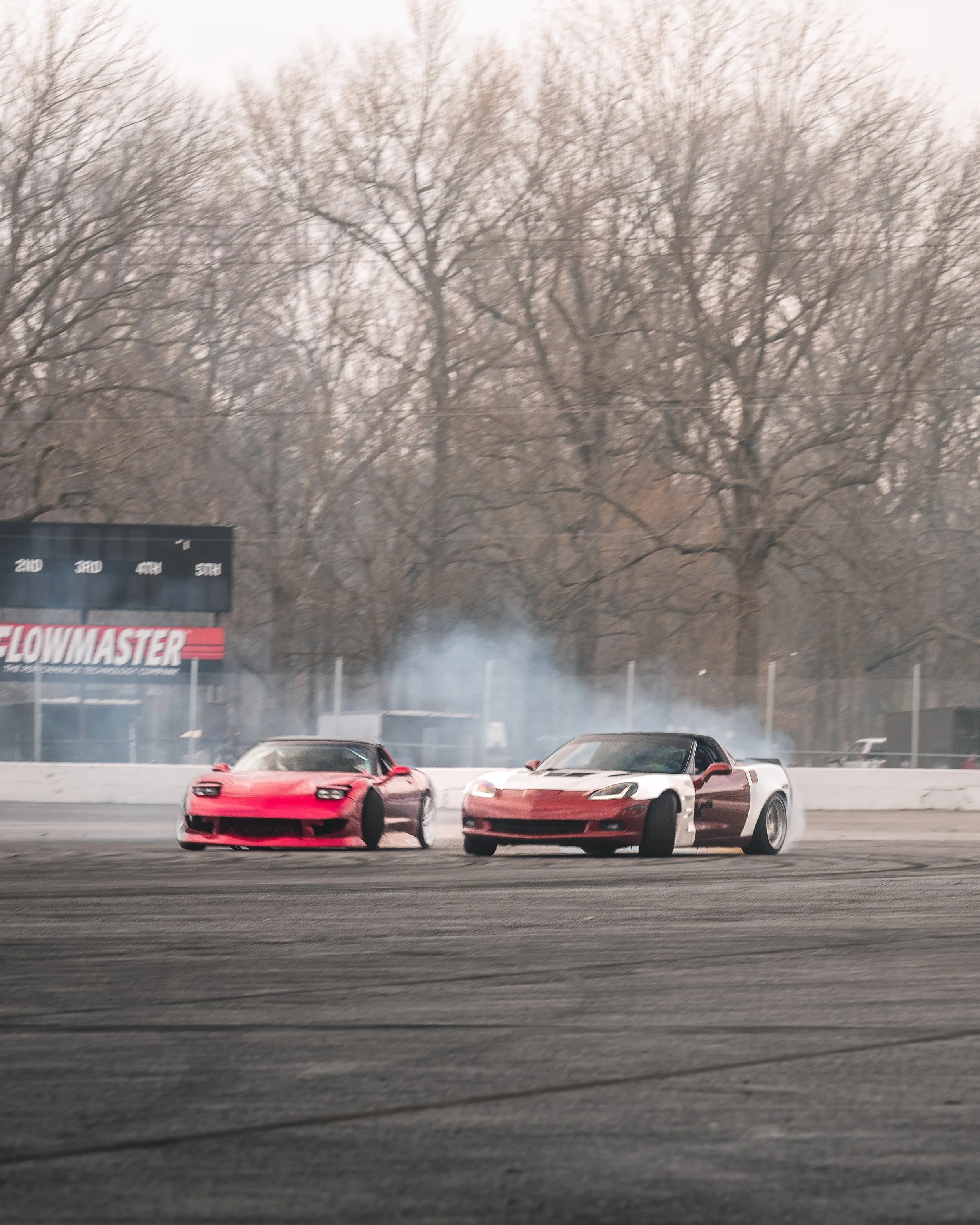 Two race cars on a track drifting with smoke in the background, trees without leaves, and a billboard reading 'Flowmaster'.