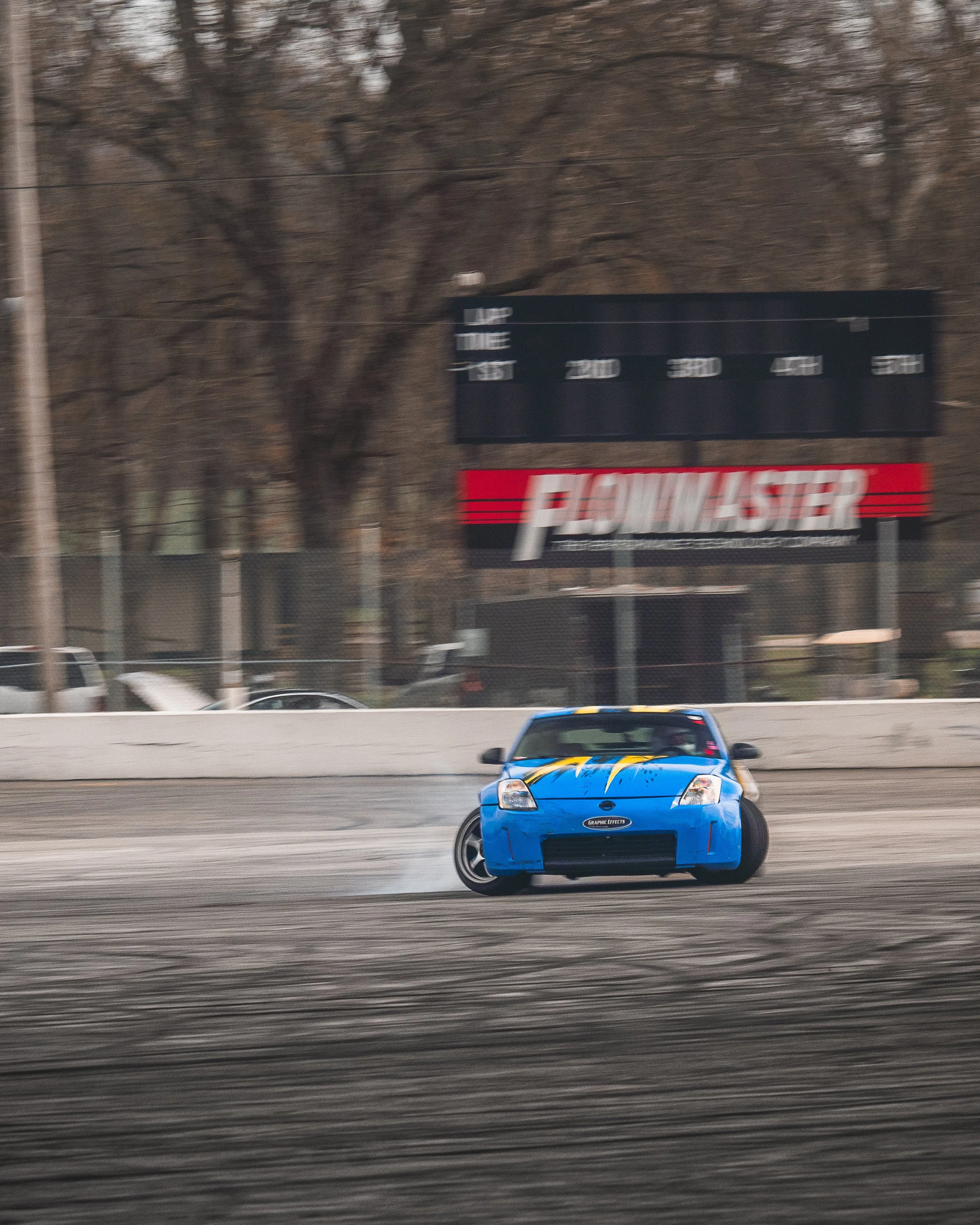 A blue race car skidding on a track, with black smoke coming from its tires, in a racing environment with a fence and trees in the background.