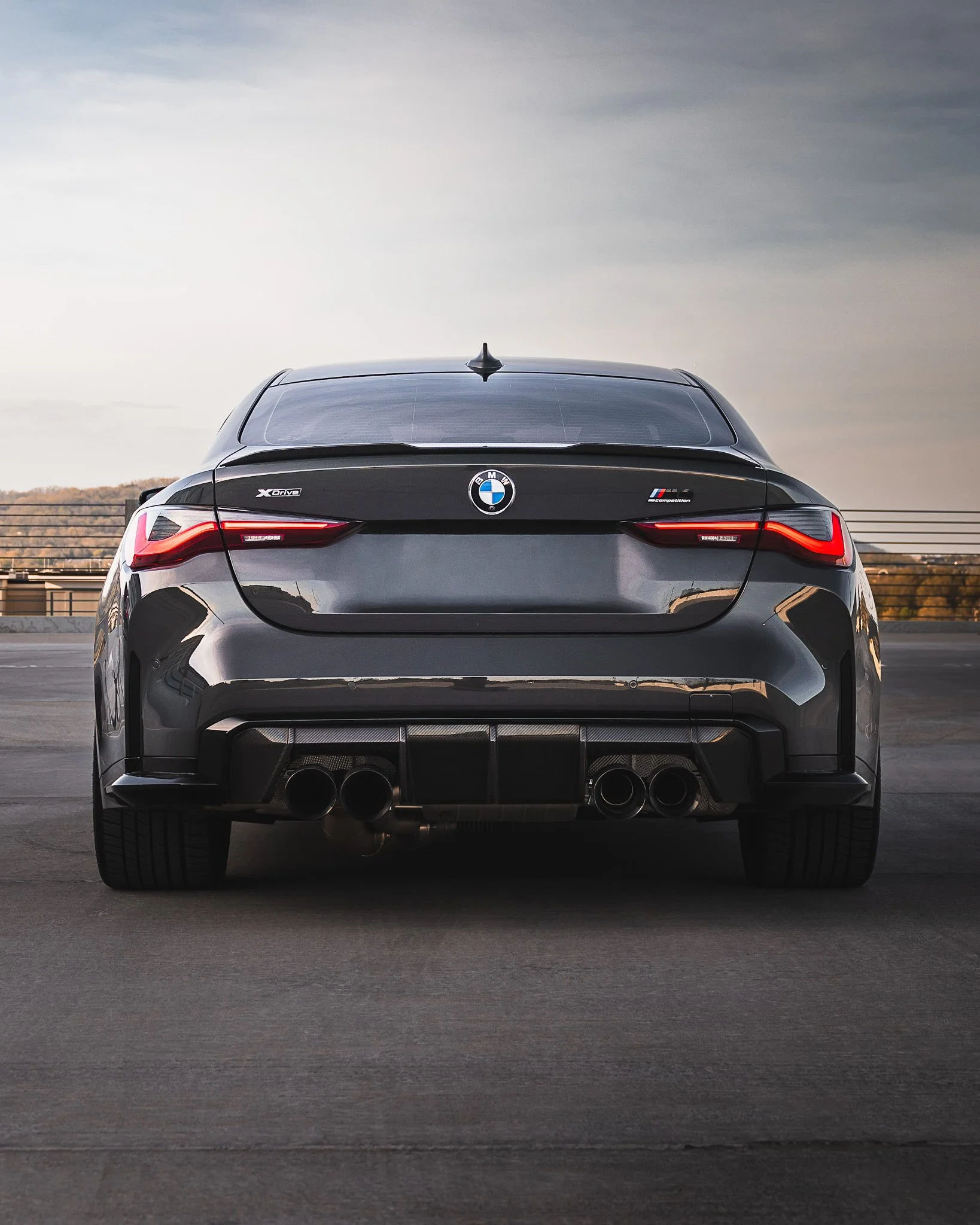 Rear view of a black BMW M car with quad exhaust pipes, parked outdoors with a cloudy sky and distant trees in the background.