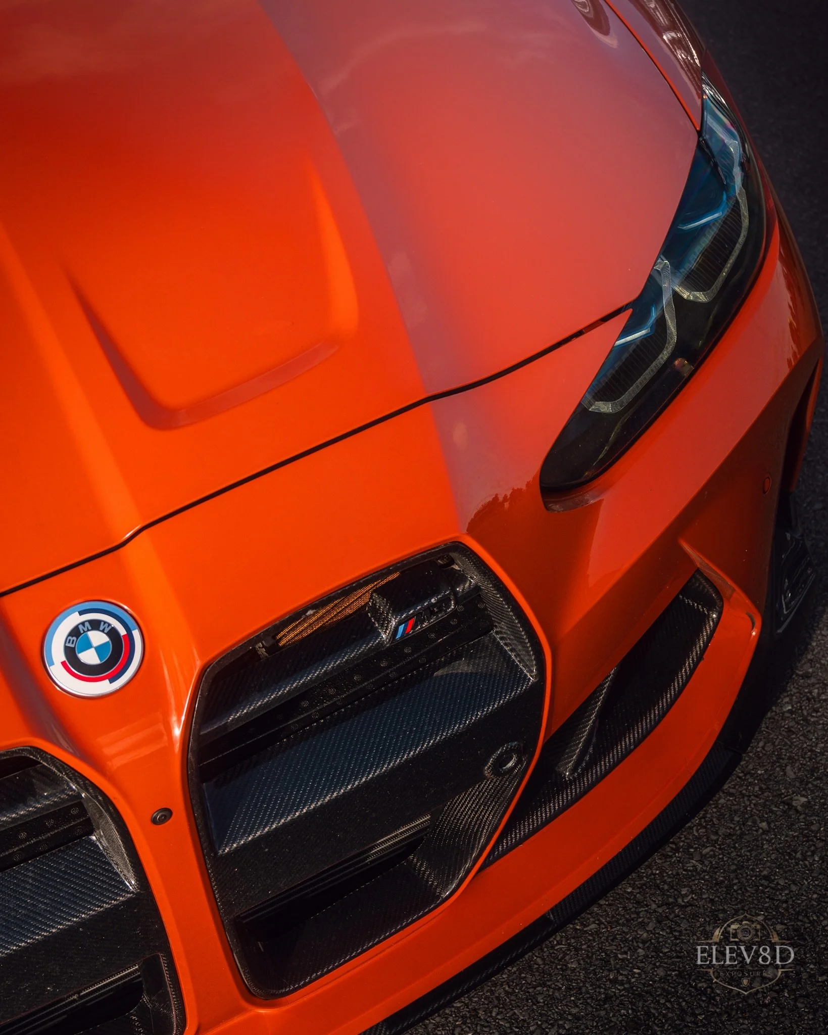 Close-up of the front of an orange BMW M car showing the BMW logo, headlights, and carbon fiber accents.