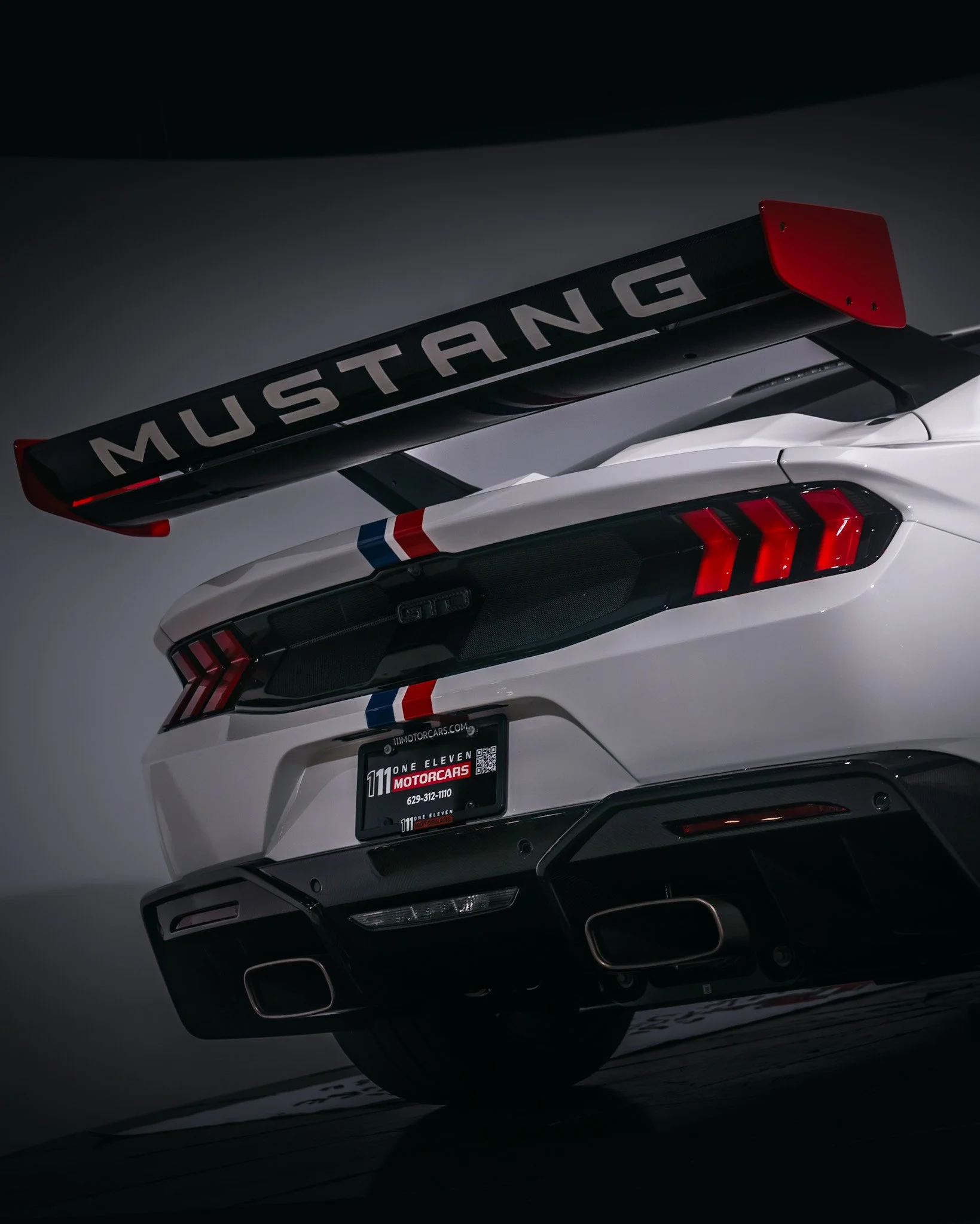 Rear view of a white Ford Mustang Shelby GT500 with racing stripes, a large rear wing with 'MUSTANG' written on it, and a dealership license plate frame.