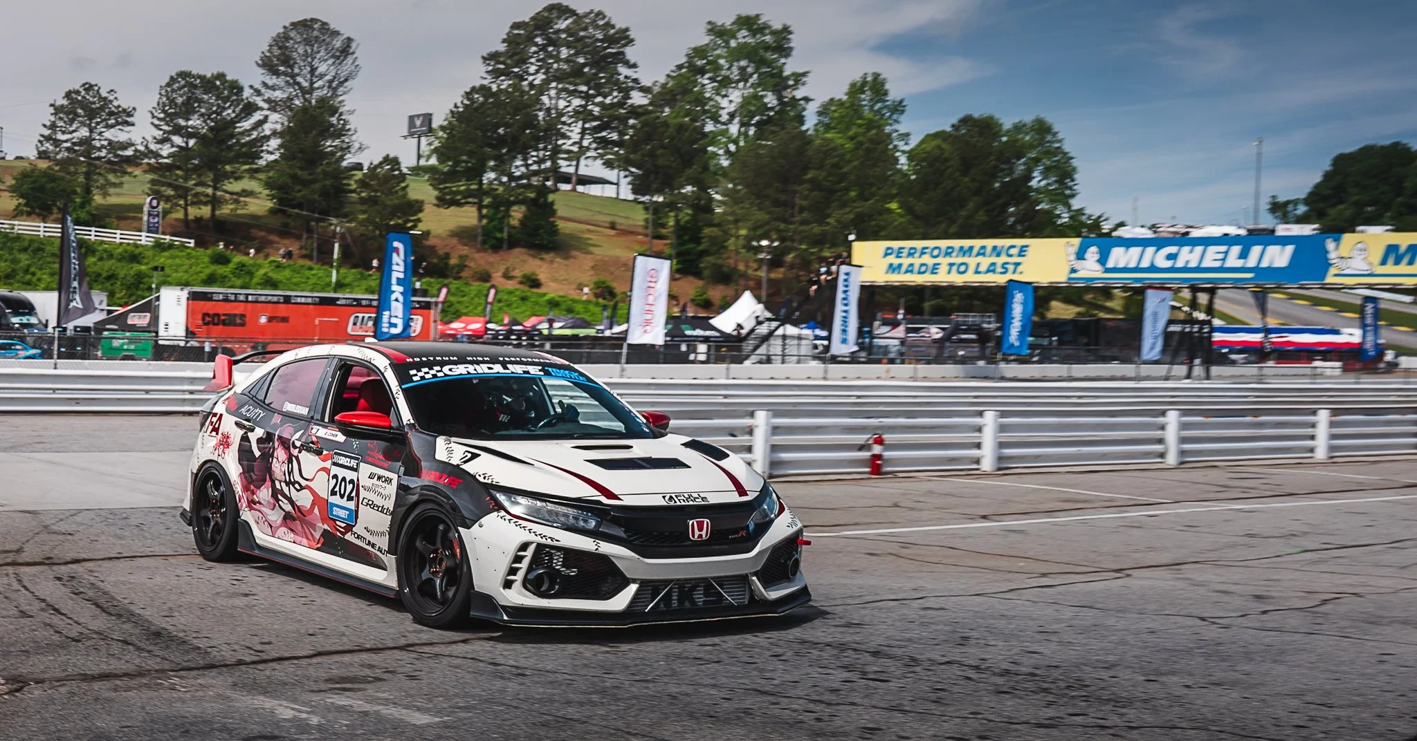 A white and black Honda racing car with red accents driving on a race track, with banners from Michelin and other sponsors in the background.