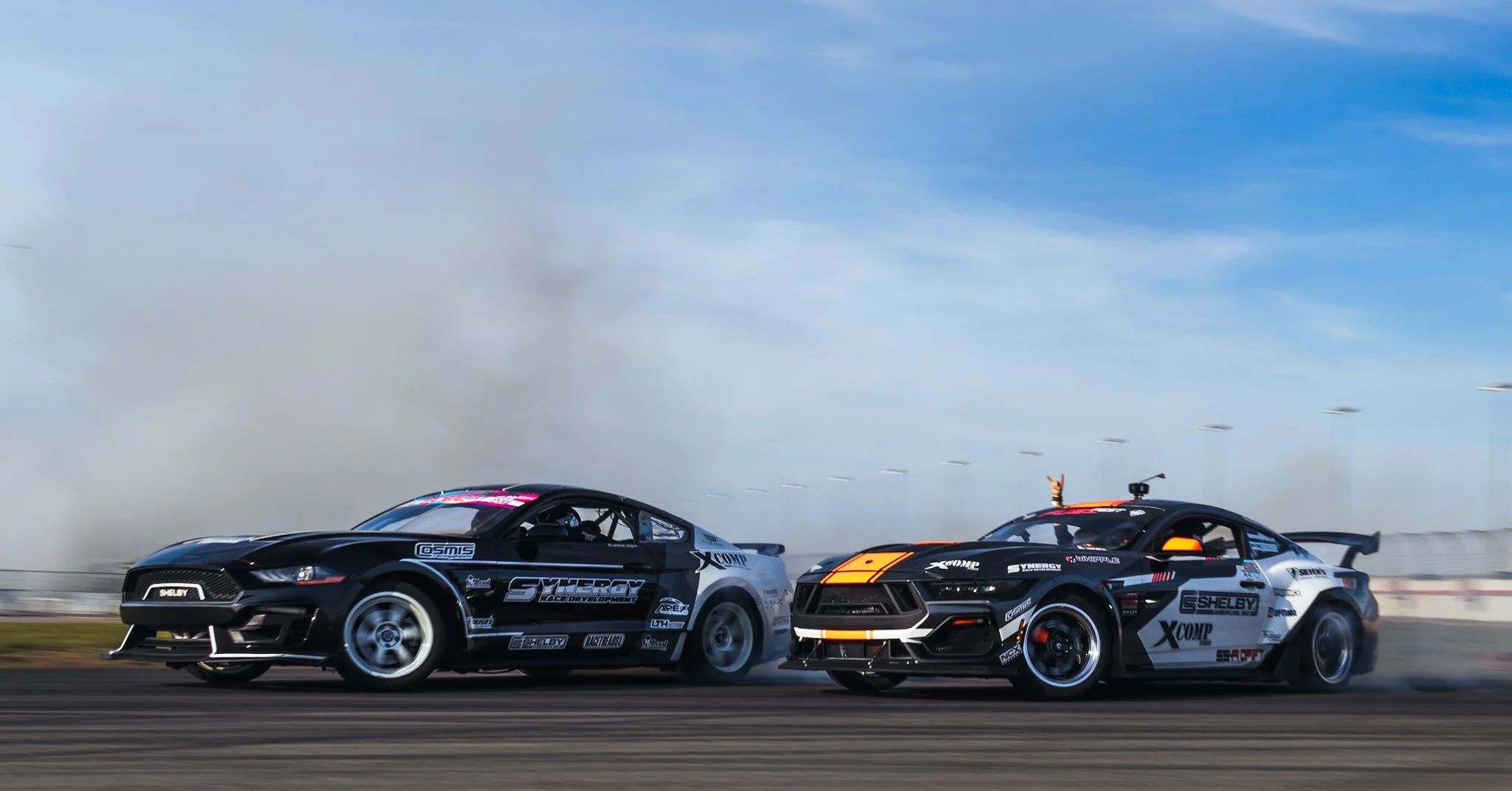 Two racing cars on a track, drifting with smoke in the background, under a blue sky, one black and the other black with orange accents.