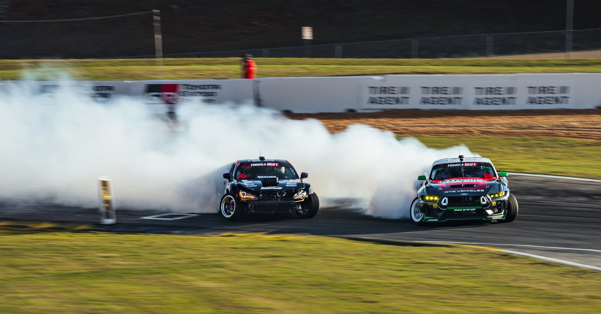 Two drift cars on a race track producing smoke as they slide around a curve, with tire barriers and a grassy area in the background.
