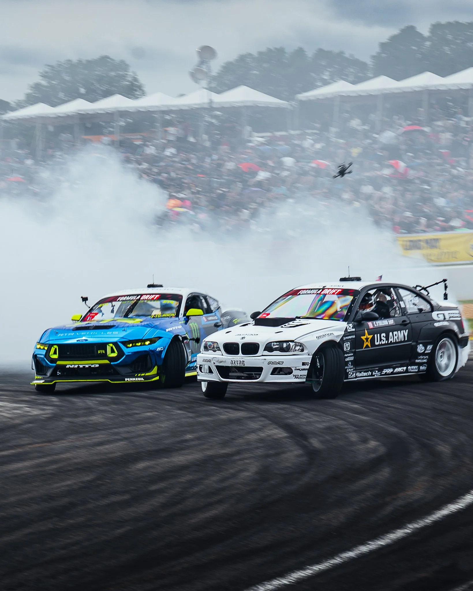 Two race cars on a track, one blue and one white with U.S. Army branding, in front of a crowd at a racing event with smoke and blurred background.