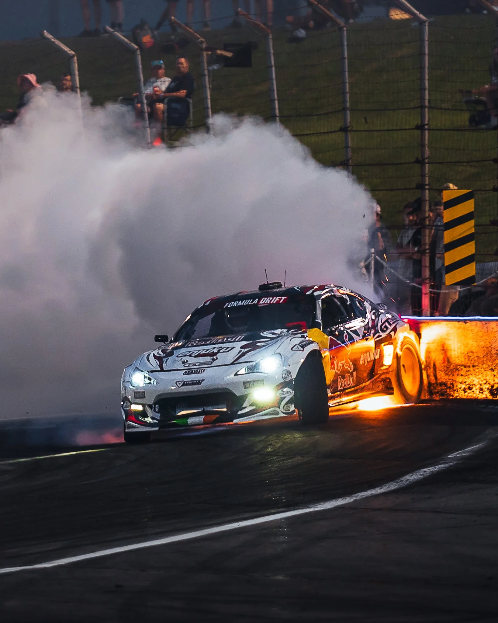 A race car, decorated with Red Bull branding, speeds along a track at night, kicking up a large cloud of smoke or dust behind it. In the background, spectators watch from behind a fence. Formula Drift Indy
