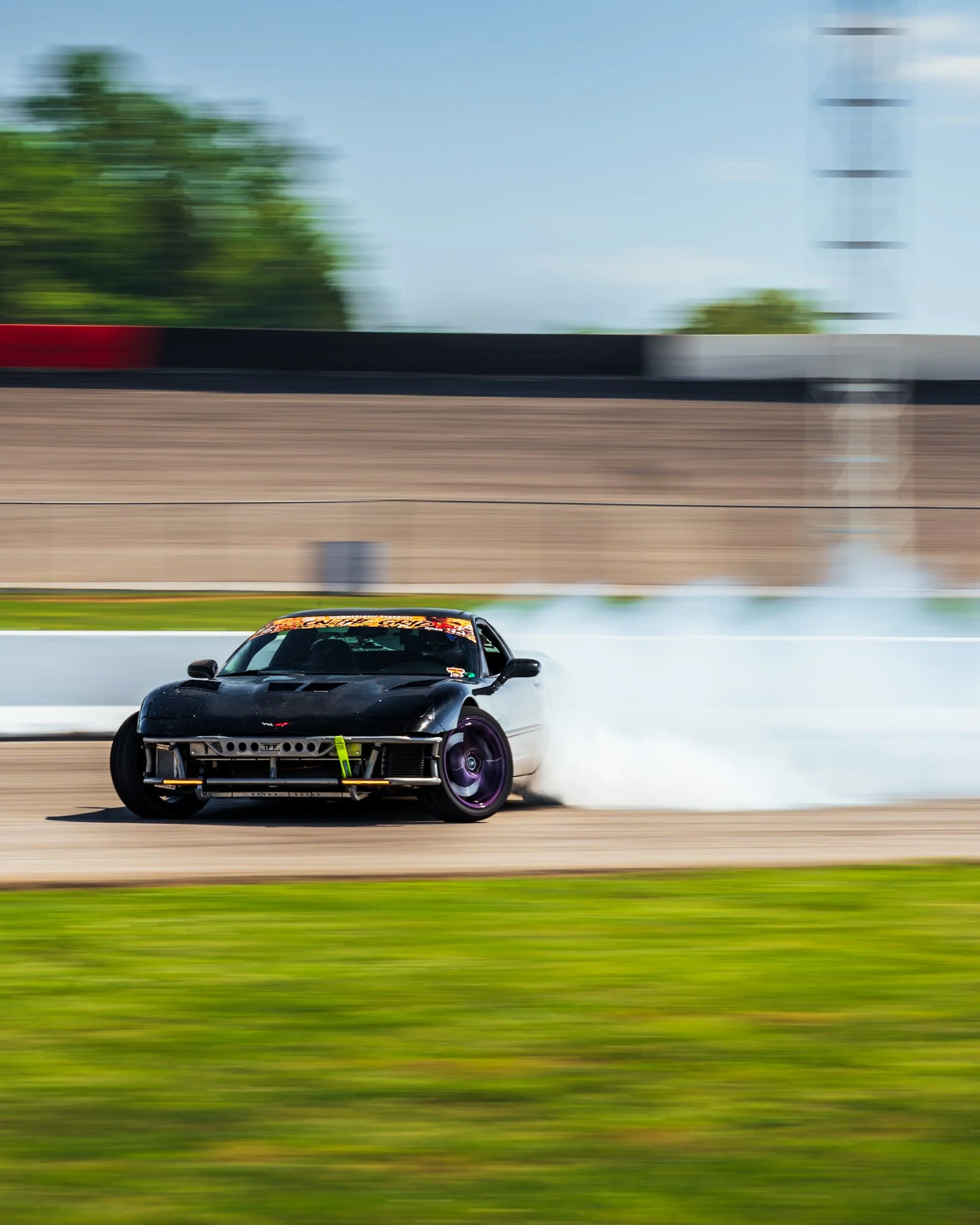 A black sports car with a yellow tow strap on the front, driving on a racetrack with a blurred background indicating high speed.