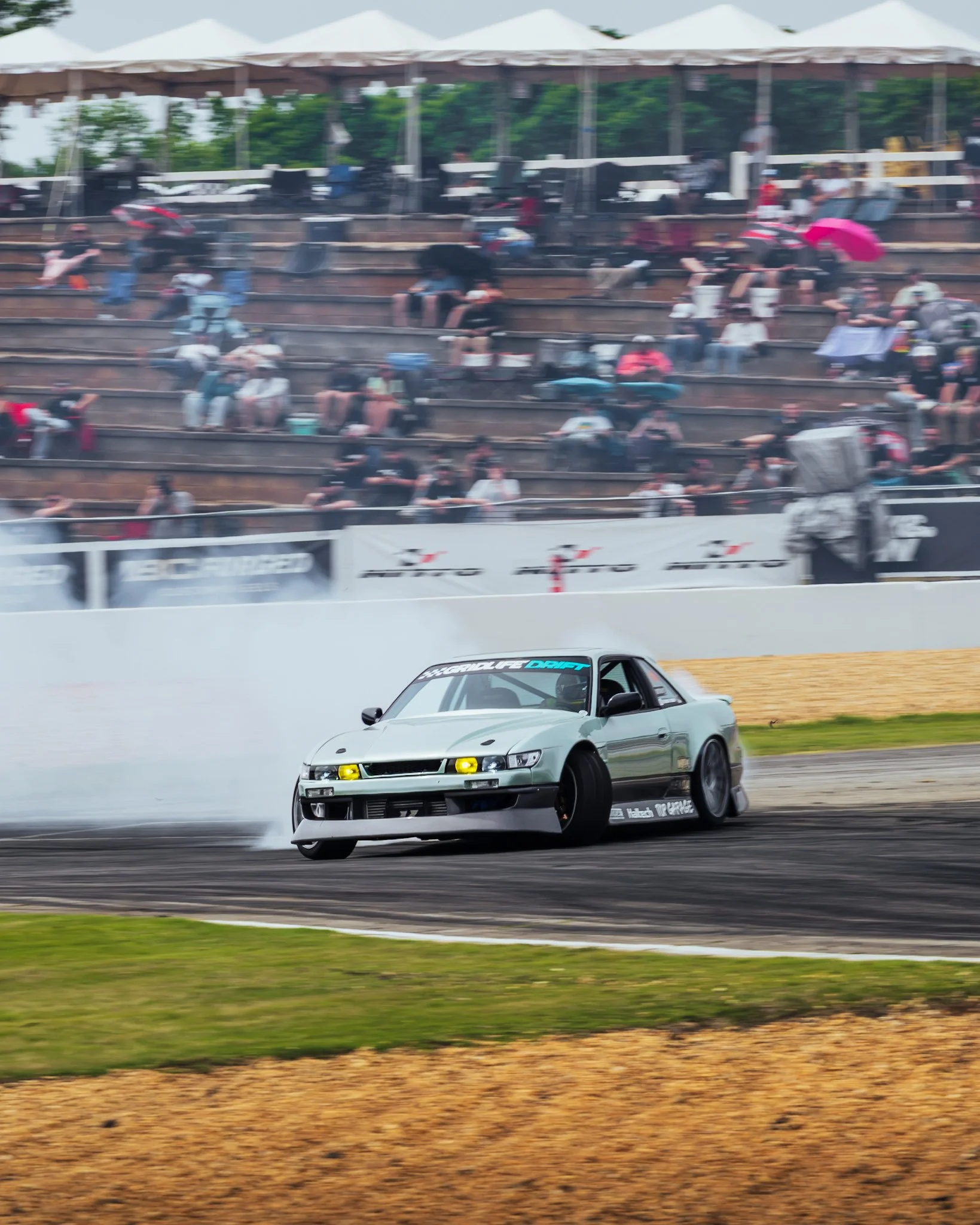 A car drifting on a race track with smoke and tire marks, while spectators sit on bleachers and watch.