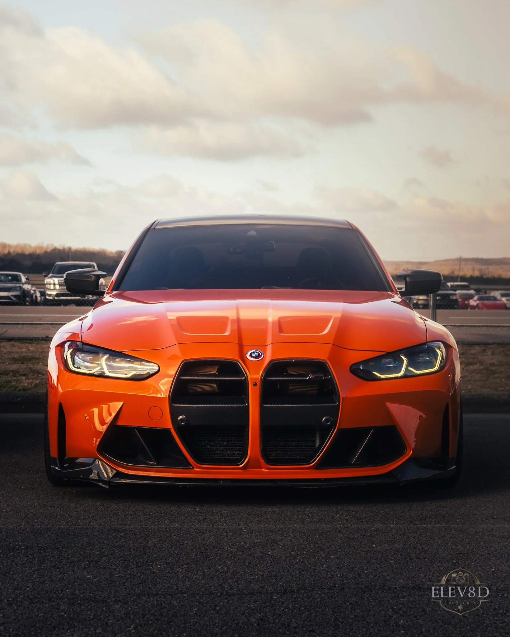 Front view of an orange BMW M3 sports car parked outdoors on a paved surface, with a background of other vehicles and a cloudy sky.