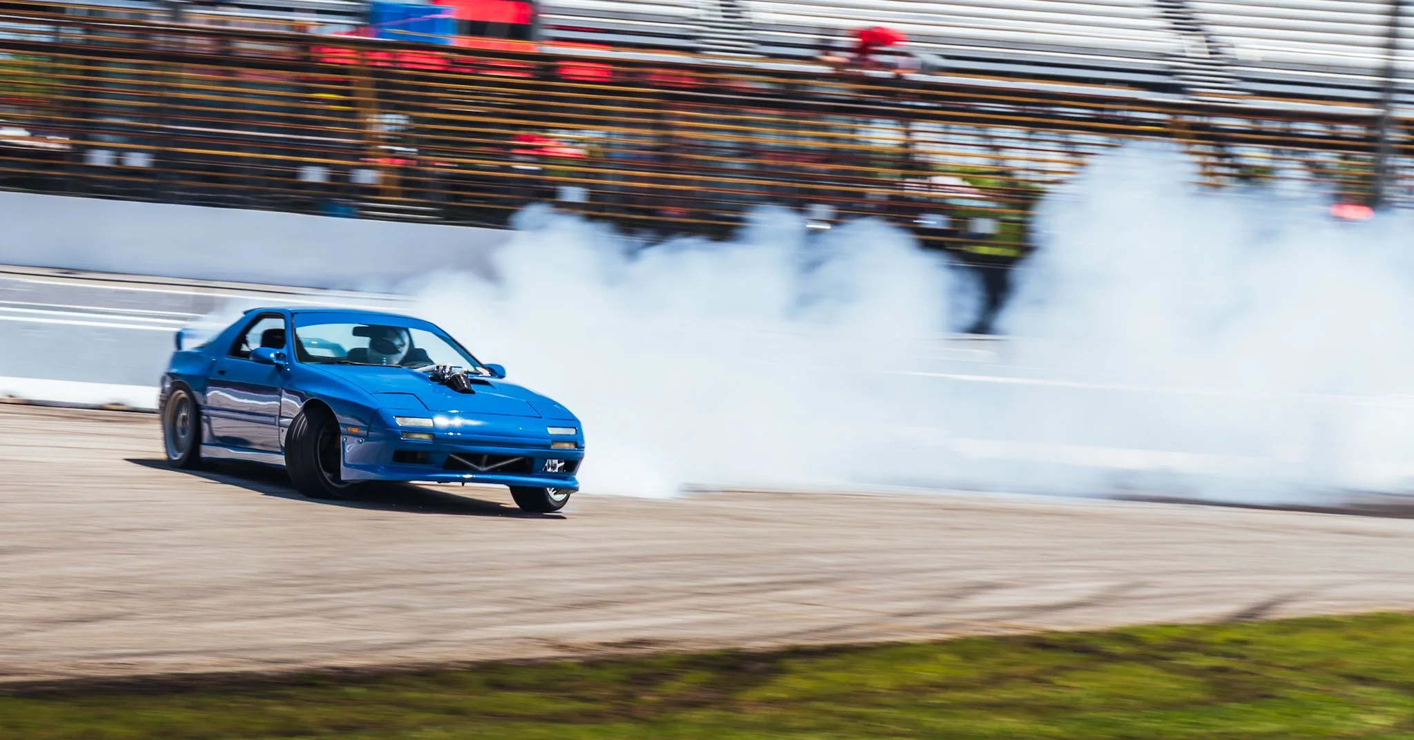 A blue sports car drifting on a race track, creating tire smoke with a background of blurred spectators and bleachers.