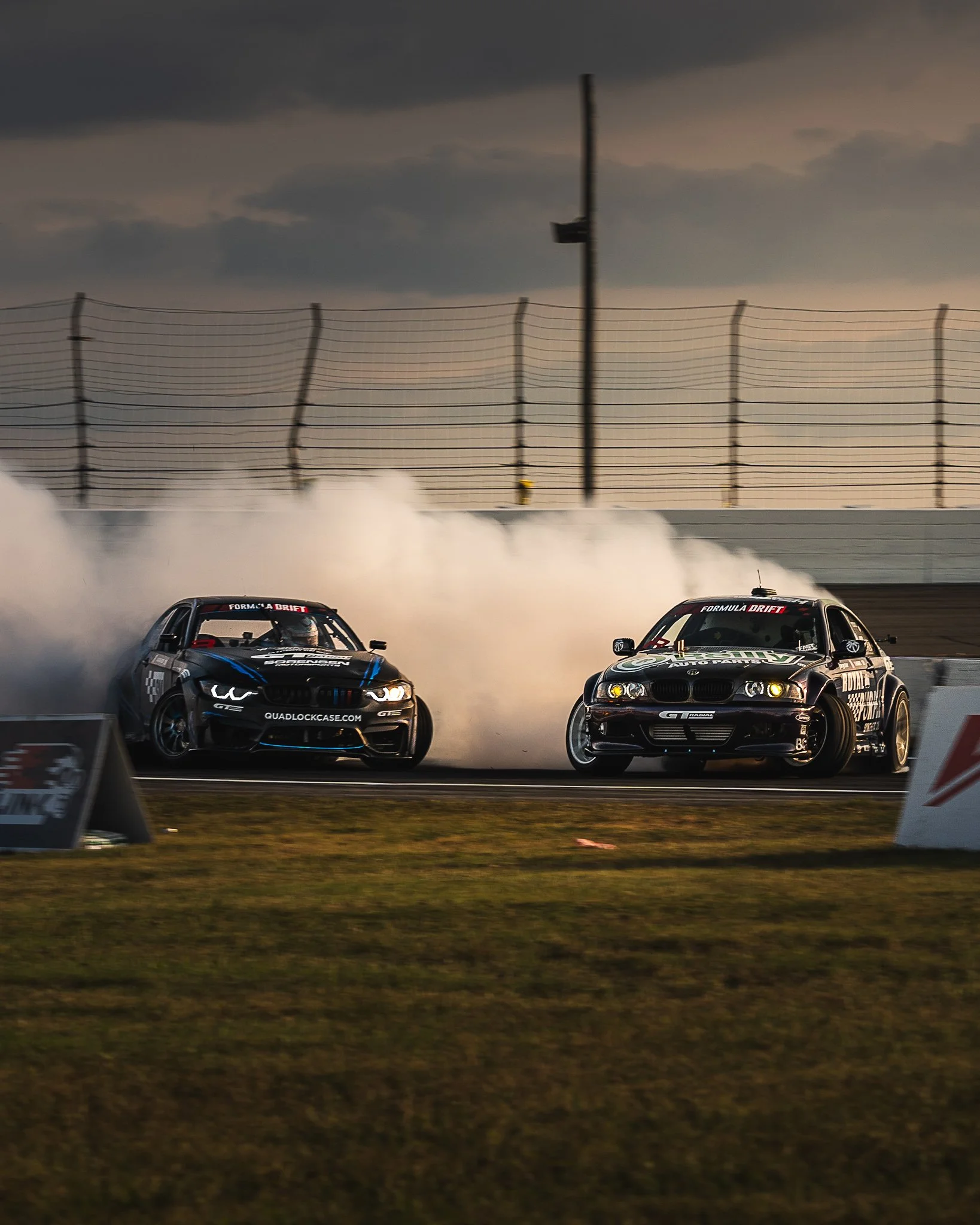 Two black race cars drifting on a race track with smoke trailing behind them, under a cloudy sky, with a safety fence and a sign in the foreground. Brandon Sorenson Dylan Hughes Formula Drift Indy
