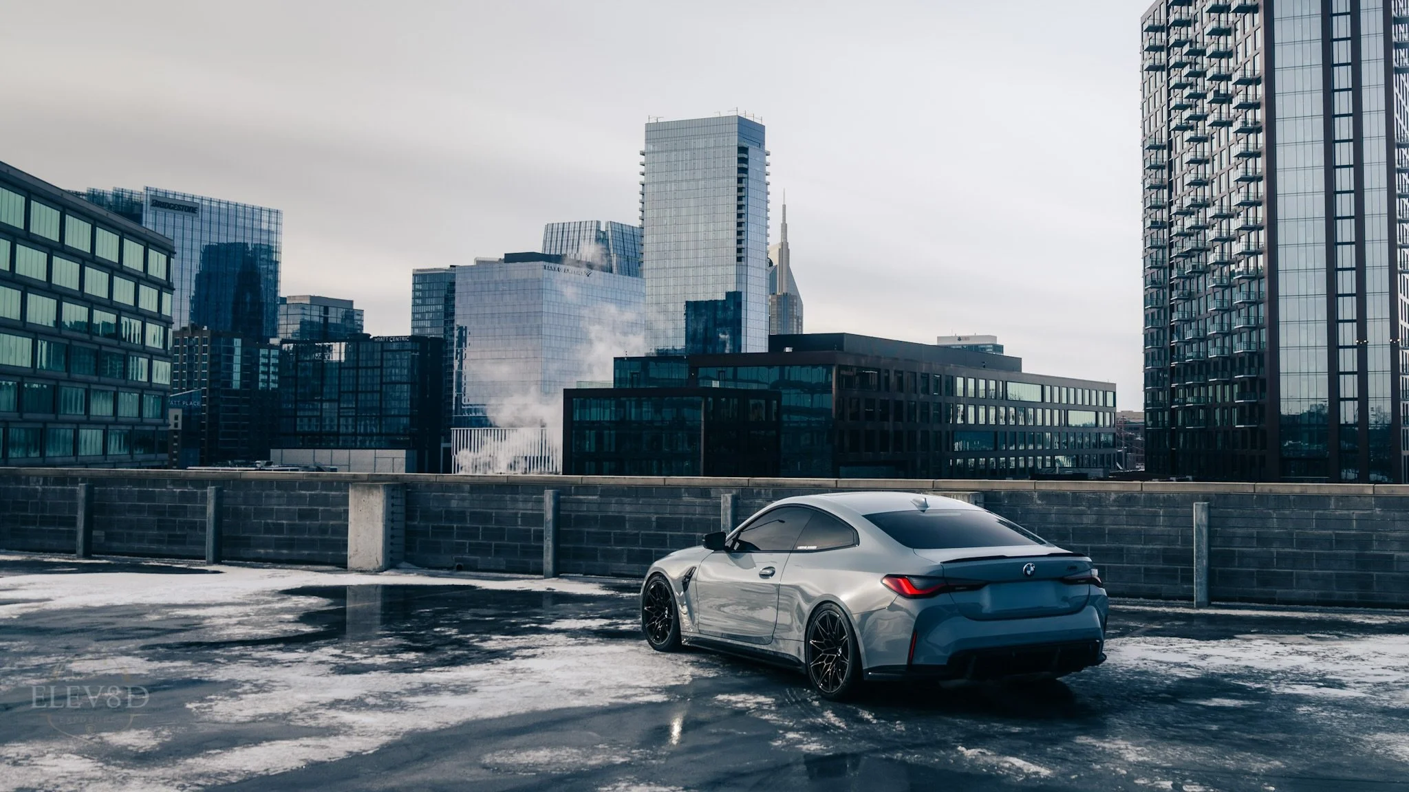 Silver sports car parked on a rooftop with a city skyline featuring tall glass buildings in the background.
