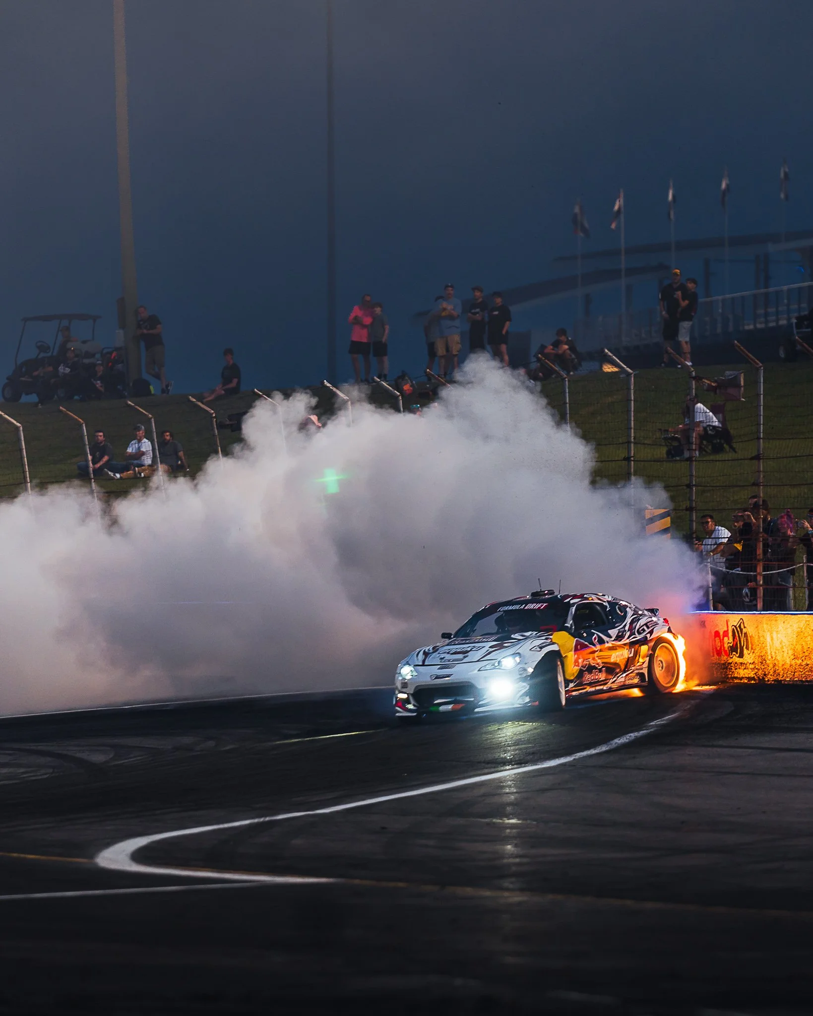 A race car drifts around a turn on a racetrack during a night race, billowing smoke as spectators watch from the stands and grassy hillside. Formula Drift Indy