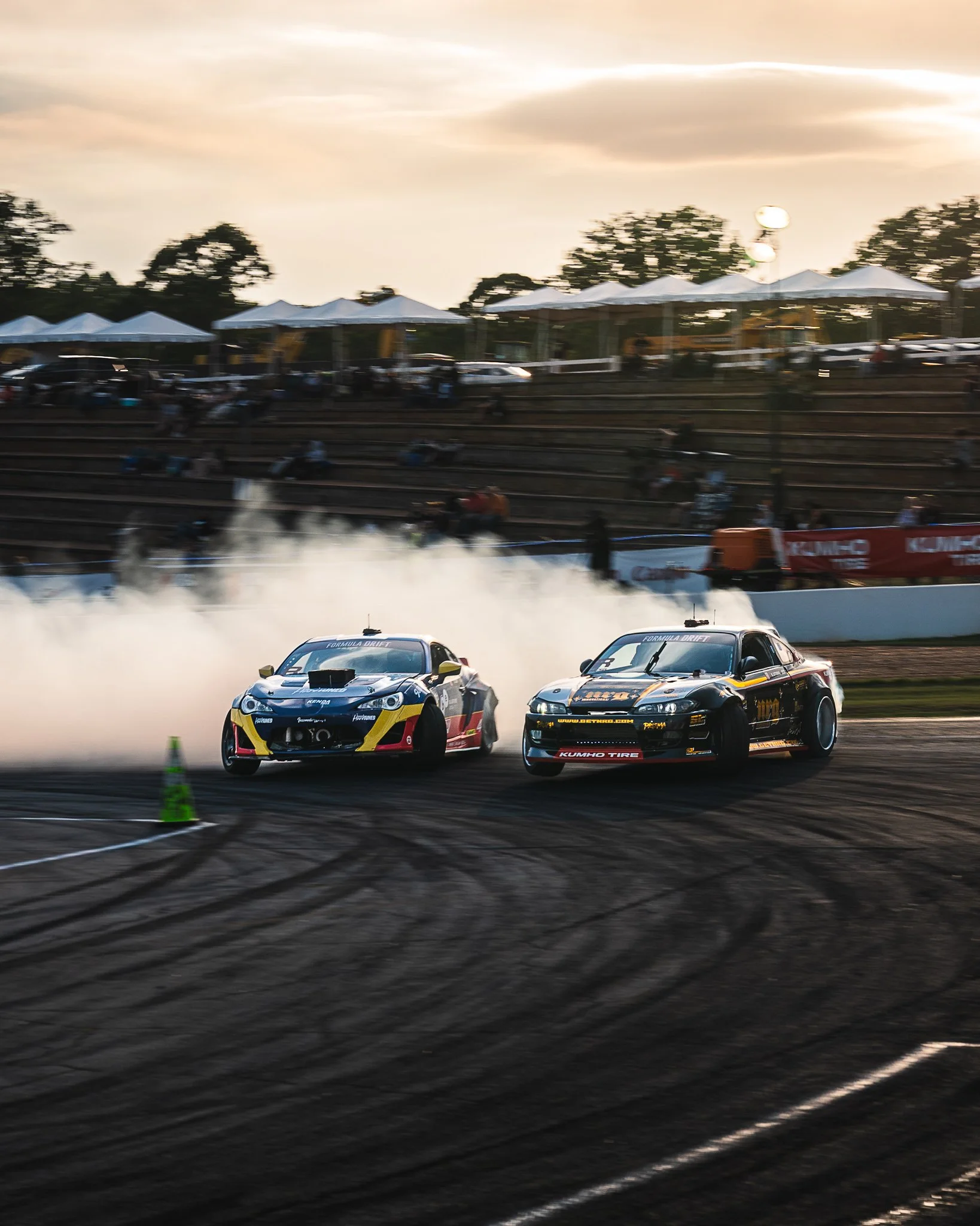 Two race cars drifting around a turn on a racetrack with smoke trailing behind. Spectators and tents in the background during sunset.
