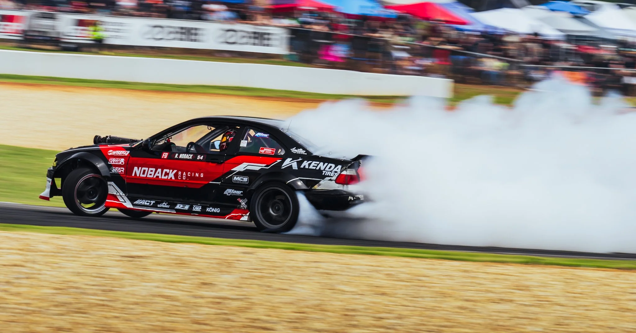 A black and red race car with sponsor logos, including Noback Racing Co., Kenda Tires, and others, drifting with tire smoke on a racetrack, with blurred spectators in the background.