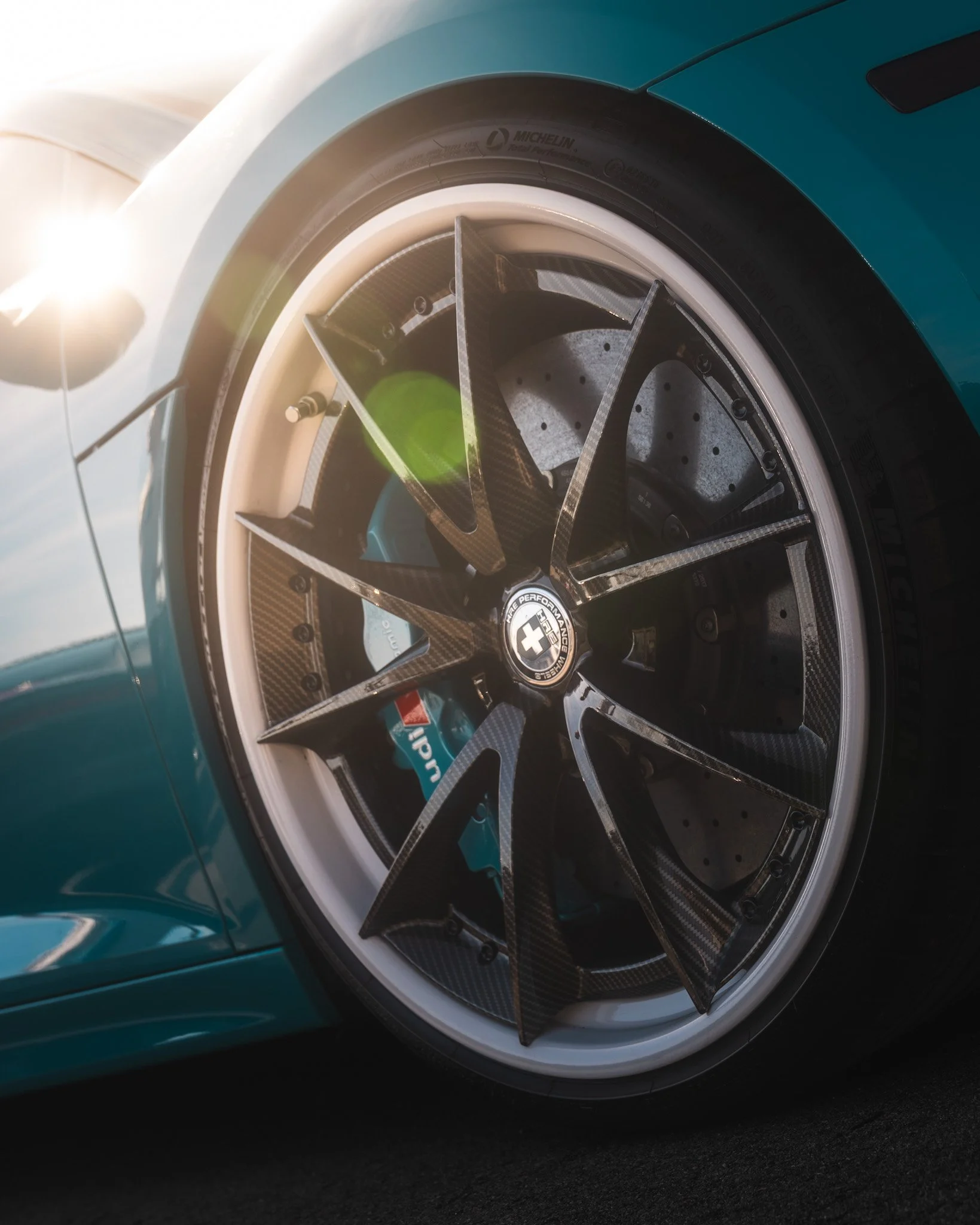 Close-up of a race car wheel with a carbon fiber rim, a high-performance brake caliper, and a low-profile tire, with sunlight glare in the background.