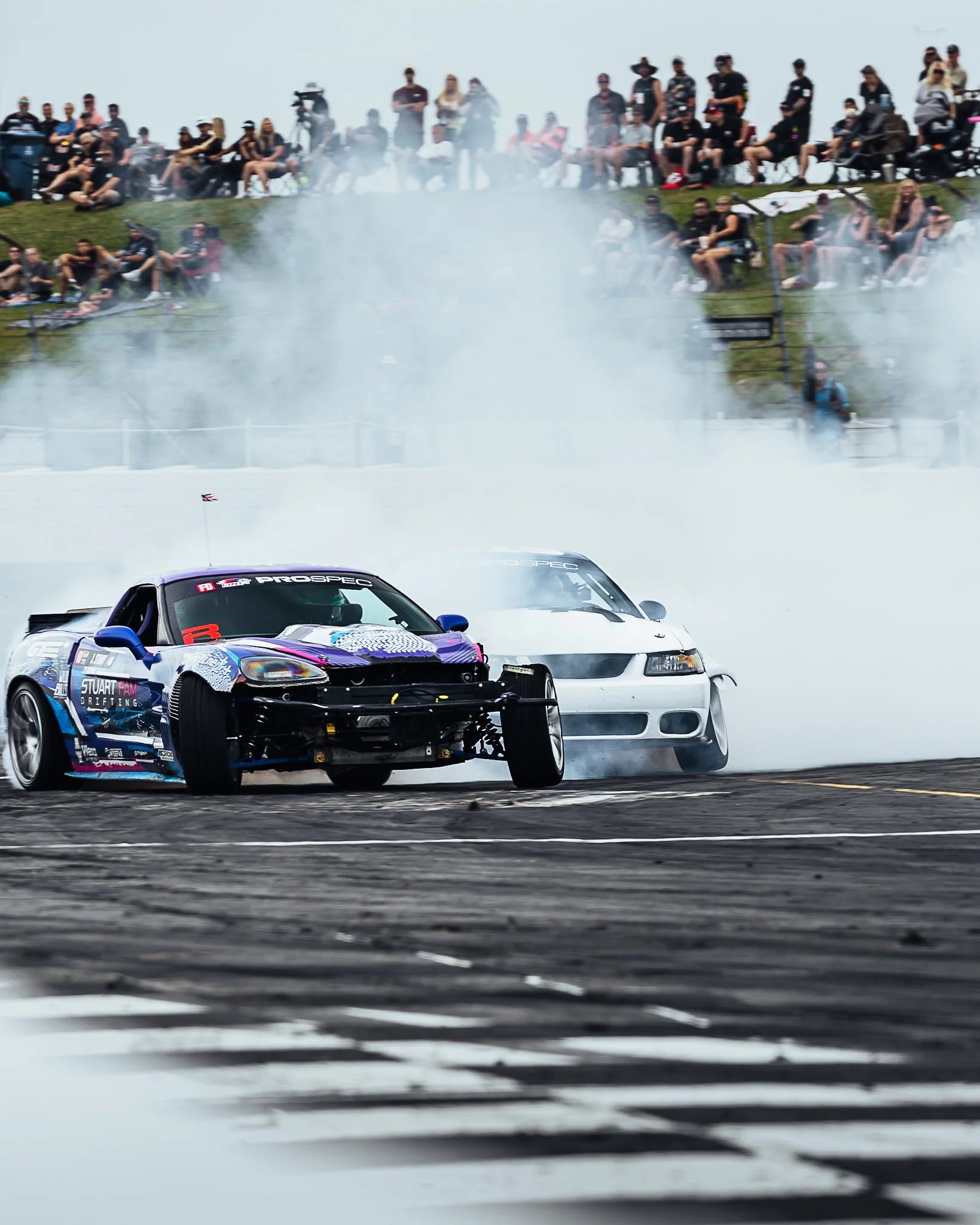 Two racing cars on a track with smoke and a crowd of spectators watching from on a hill.