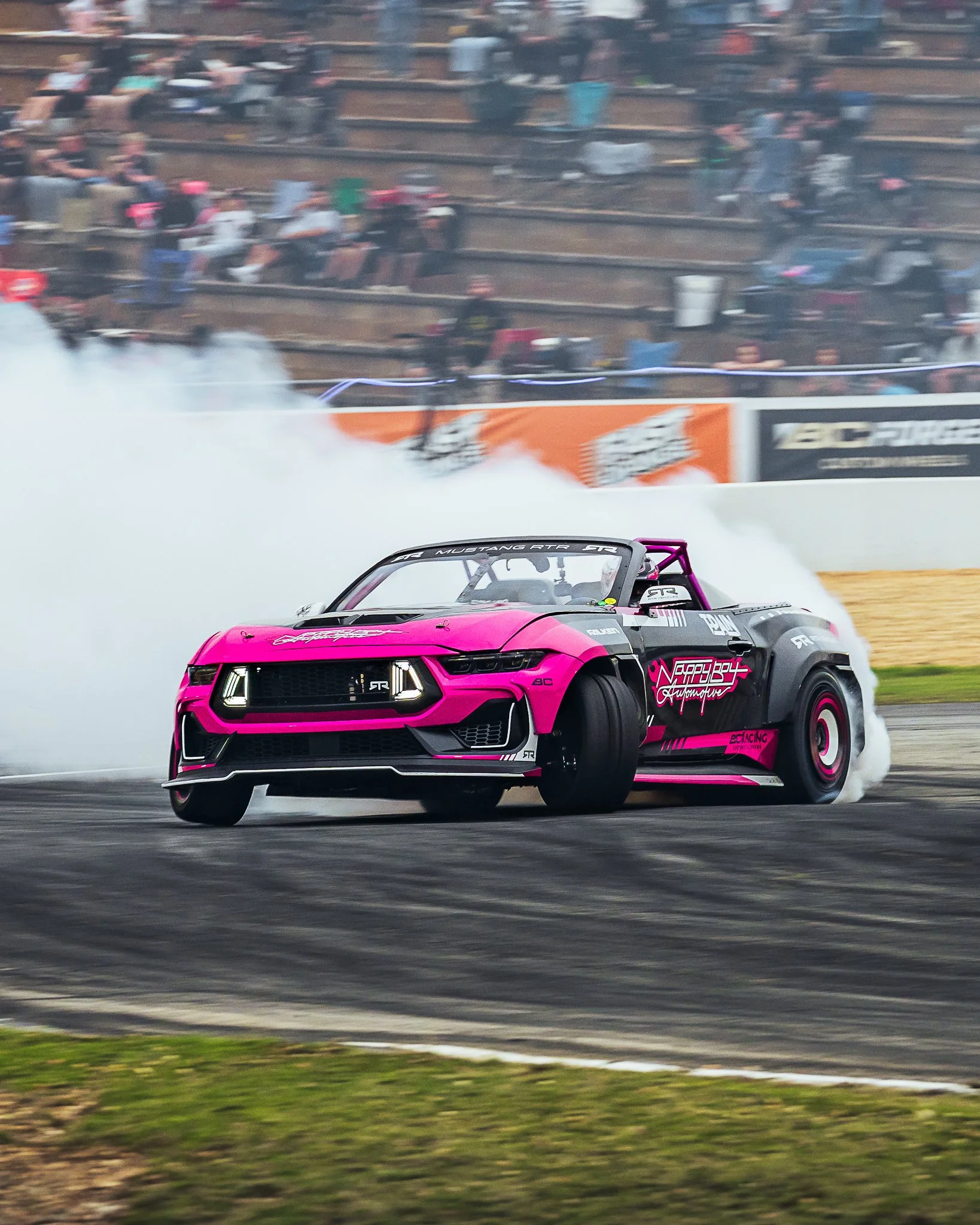 A pink and black race car drifting on a track with smoke coming from its tires, with a crowd watching in the background.
