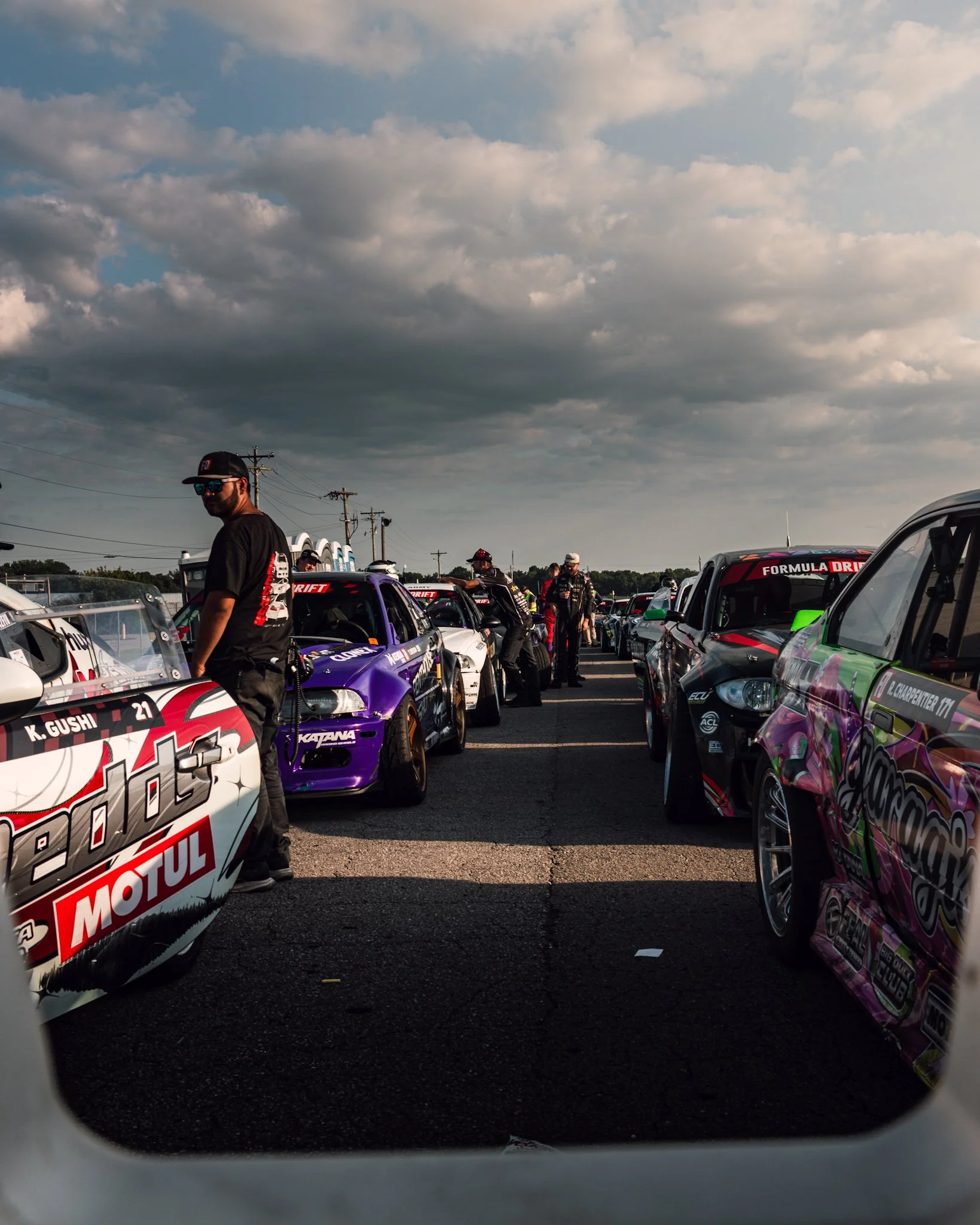 Race cars lined up in a paddock area with people walking around; a man with sunglasses and a cap looking toward the camera; cloudy sky overhead. Formula Drift Indy