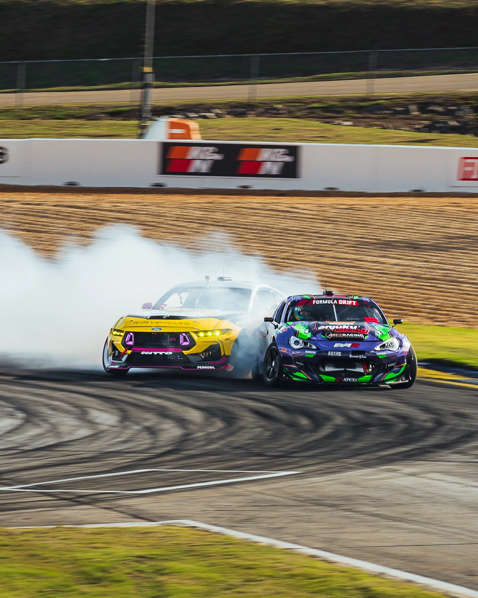 Two race cars on a track, one yellow and one black with colorful graphics, drifting with tire smoke behind them during a motorsports event.