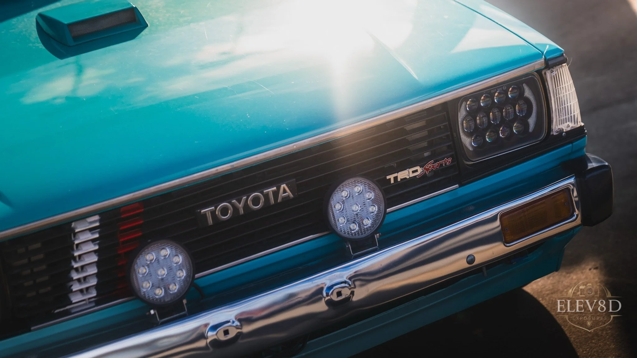 Close-up of the front of a vintage turquoise Toyota car, featuring LED headlights, Toyota emblem, TRD badge, and sporty fog lights.