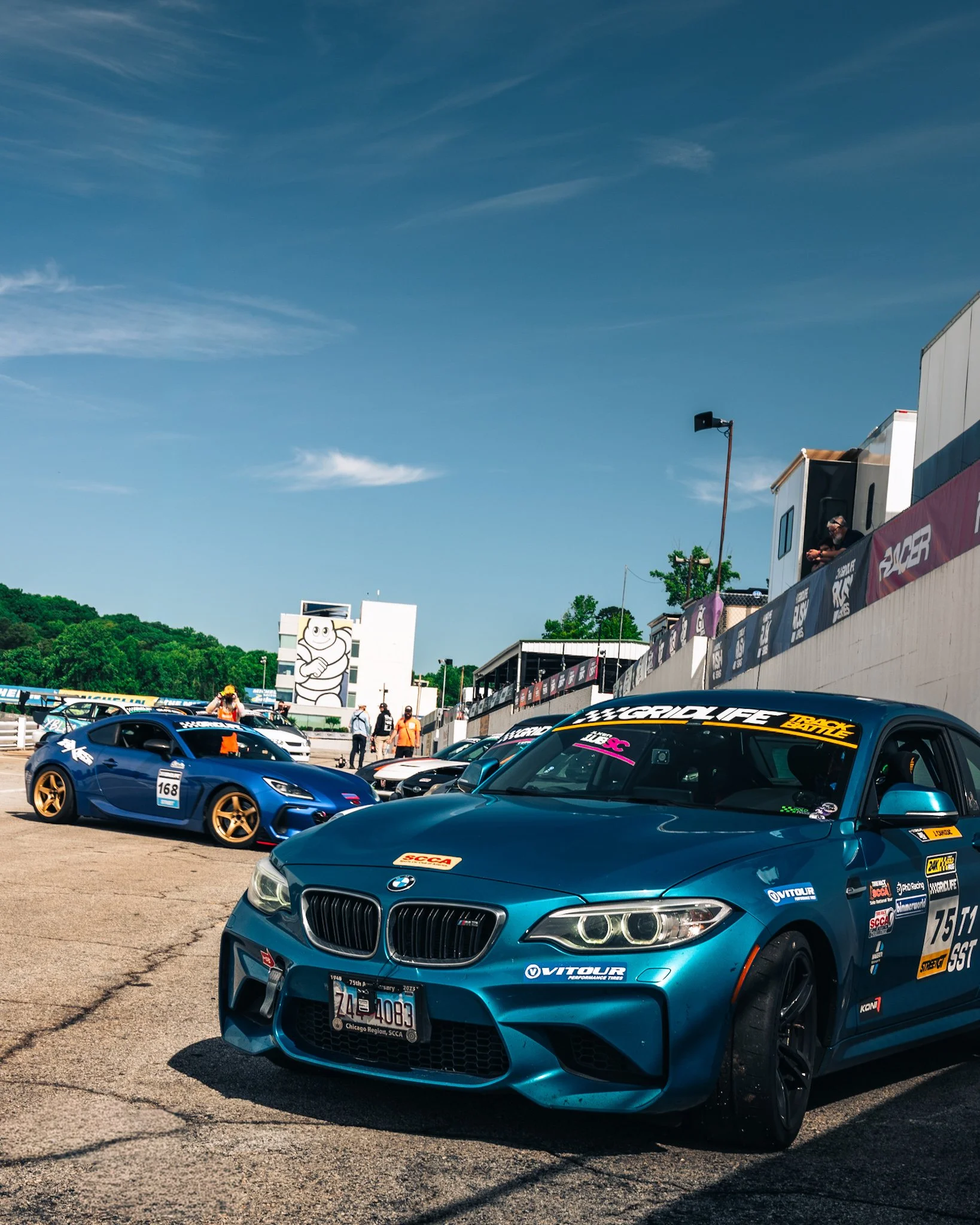 A parking lot filled with race cars, including a blue BMW and other sports cars, under a bright blue sky with some clouds. The cars have racing decals and stickers, and there are people in the background near a building with a cartoon character logo and a flags banner.