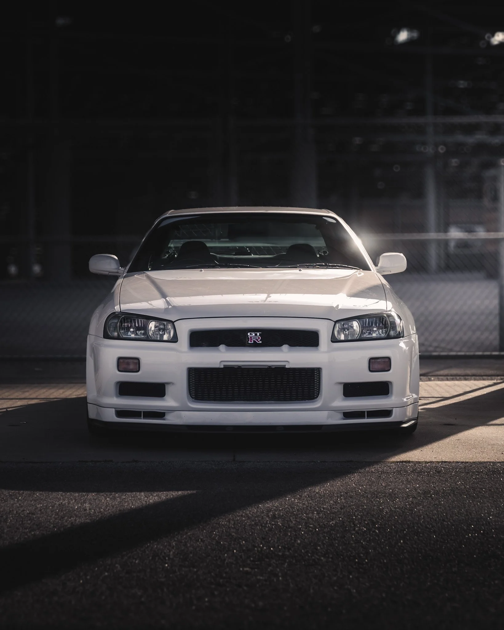 Front view of a white Nissan GT-R sports car parked on asphalt, with a dark background and sunlight reflecting off its hood.