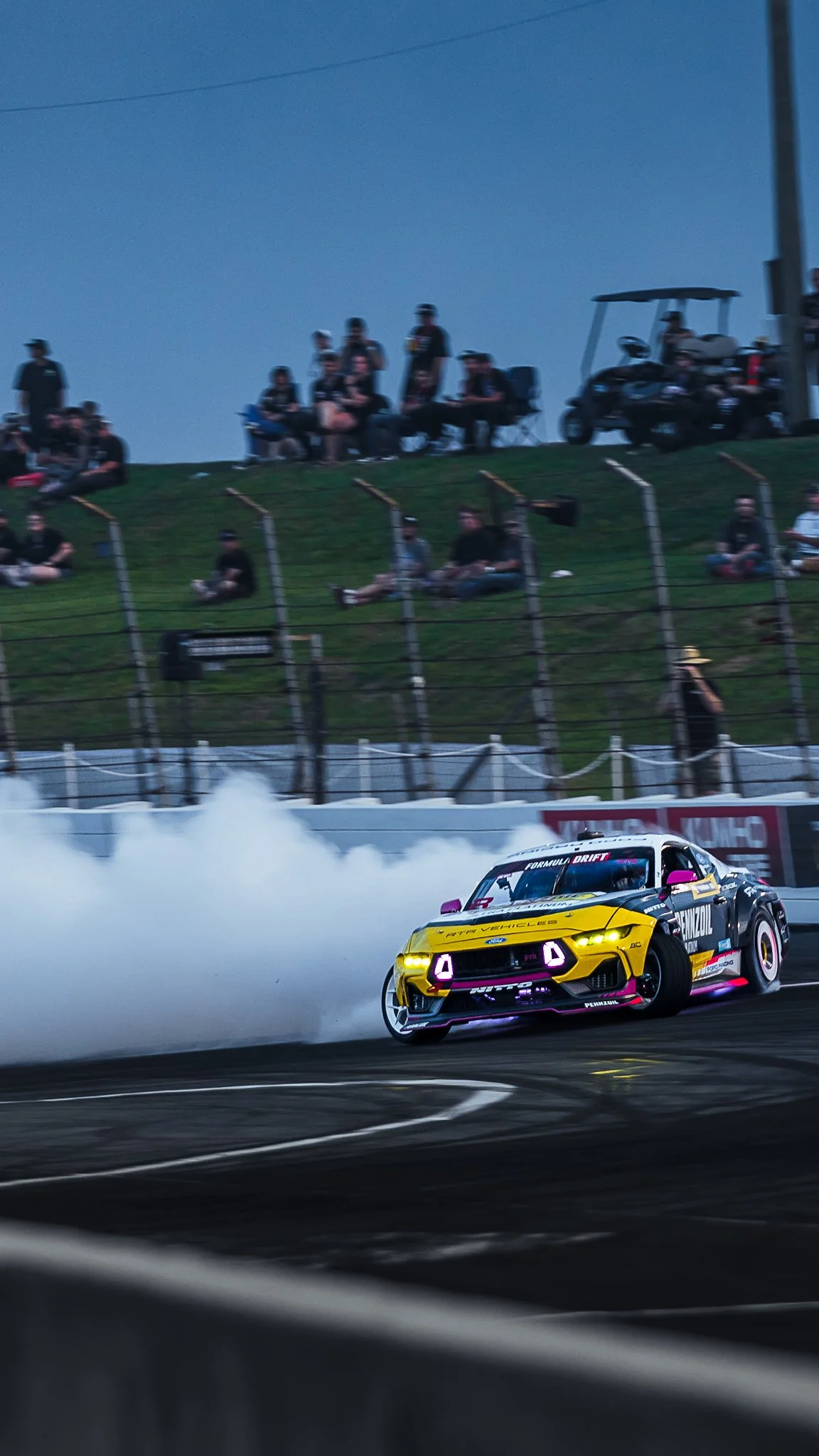 A colorful drift racing car on the track with smoke behind it, spectators watching from the hill and seating area, and a golf cart nearby at a race event during the evening. Ben Hobson Formula Drift Indy