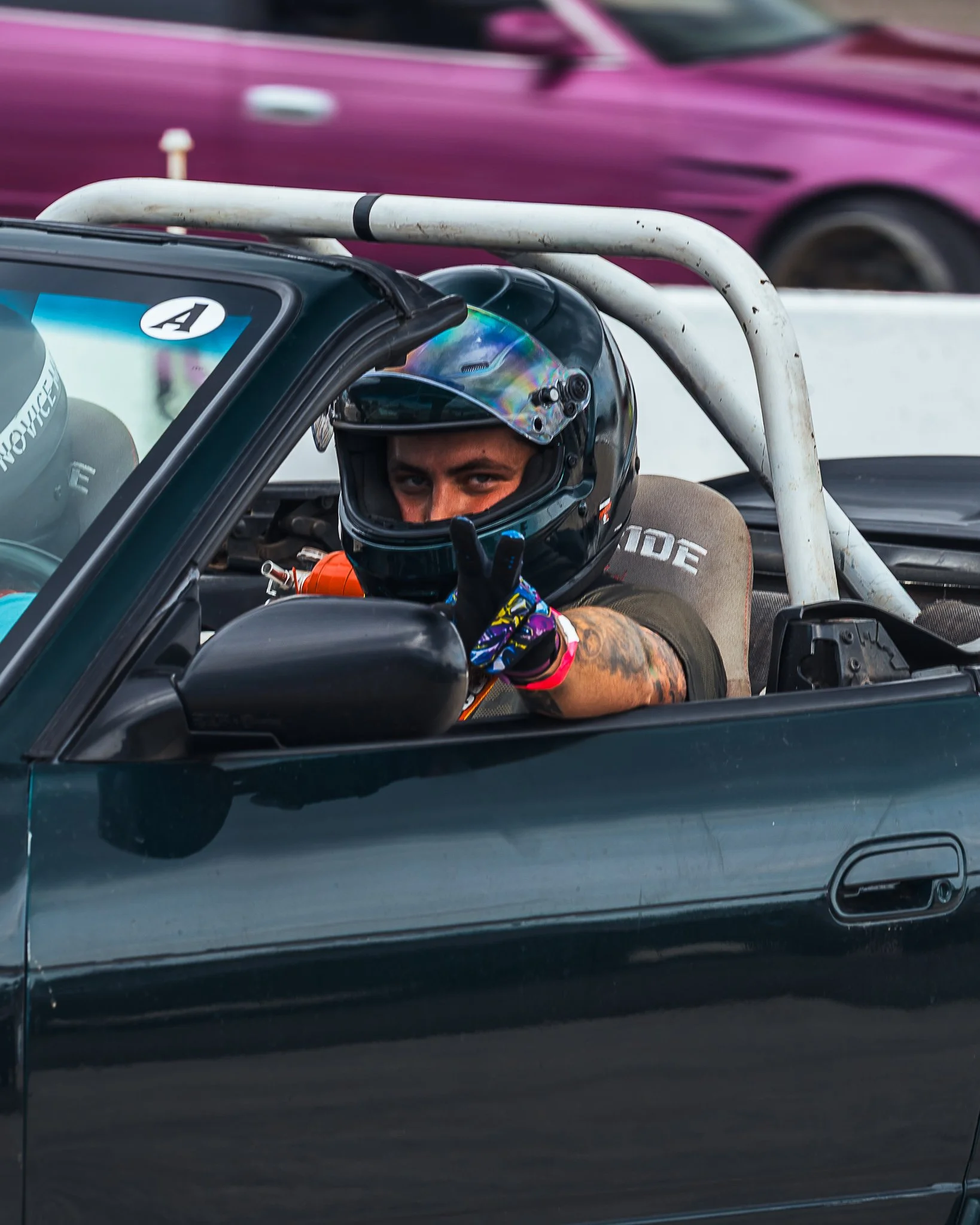 A person wearing a racing helmet inside a race car, making a peace sign with one hand, with a pink car passing by in the background.