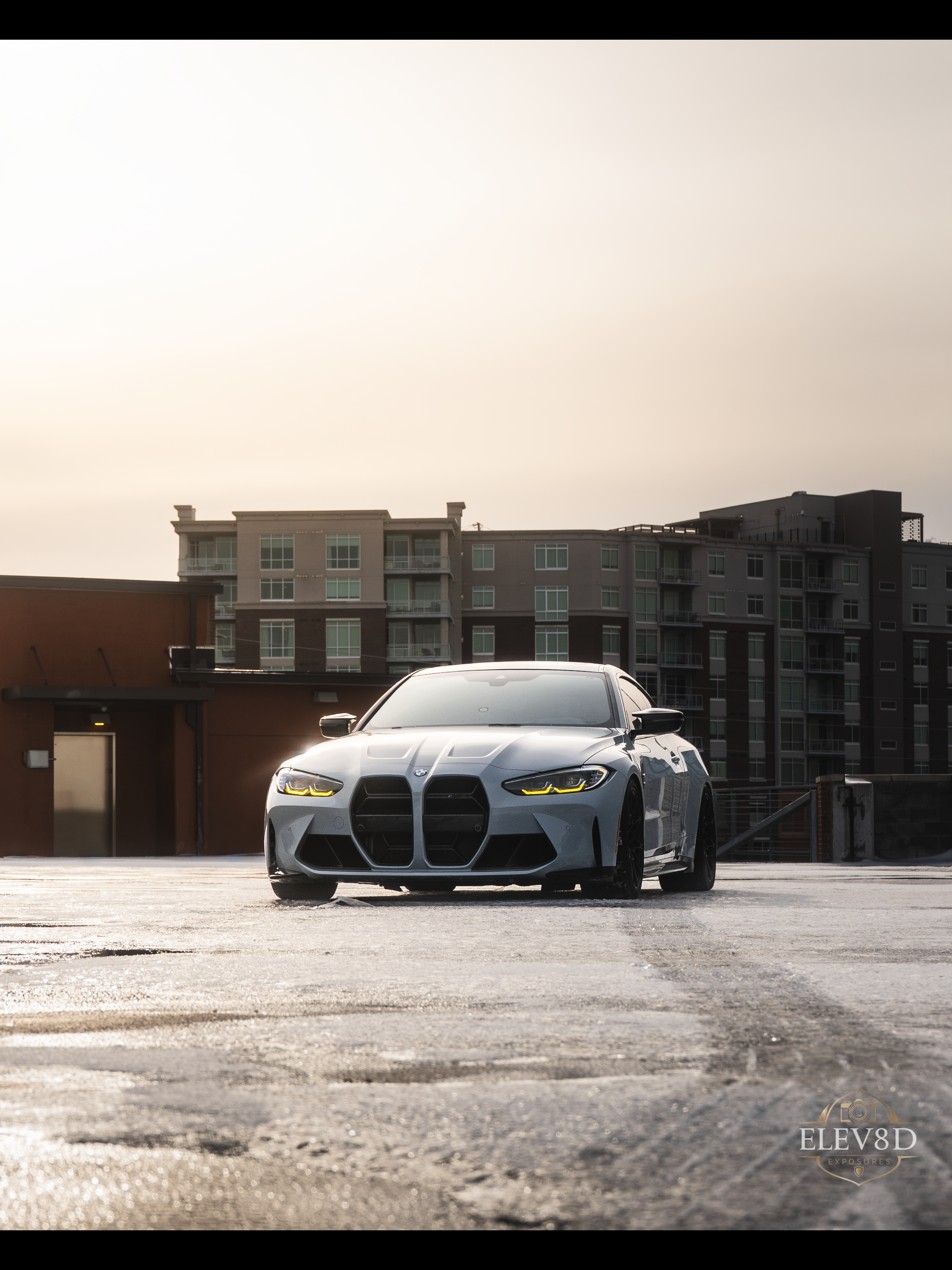 A sleek gray BMW M3 sports car parked on a wet rooftop parking lot with a city apartment building in the background and a cloudy sky overhead.