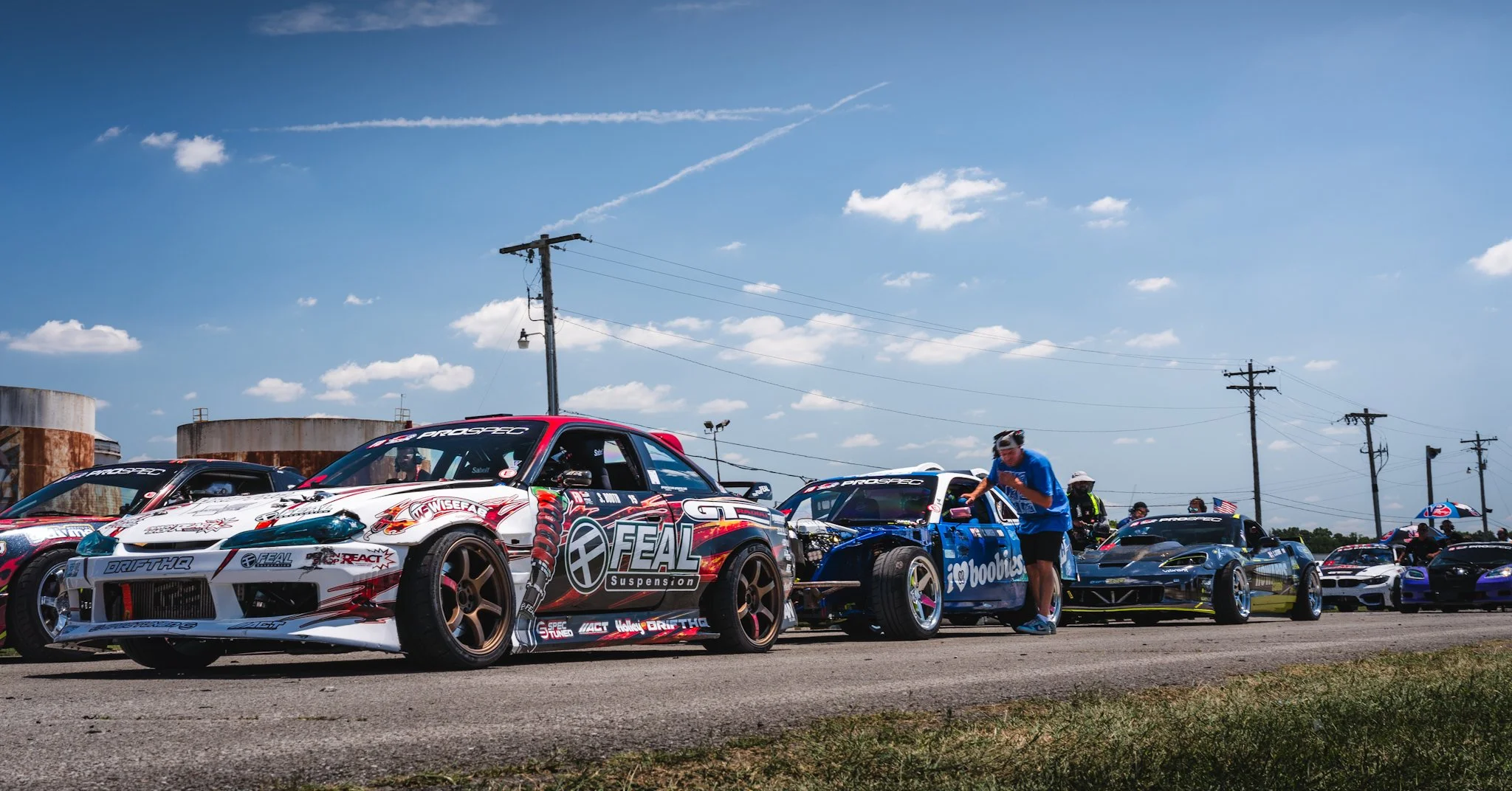Race cars parked in a line at a racing event with people inspecting and working on them, under a blue sky with clouds and power lines in the background.
