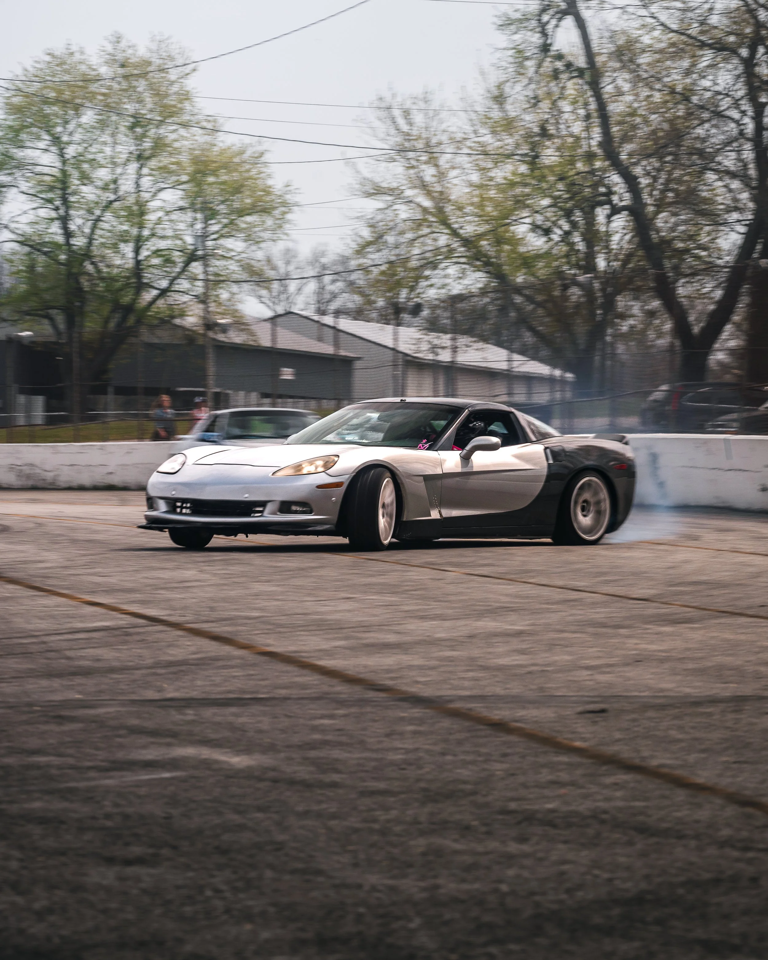 A silver and black sports car drifting on a race track, creating smoke from the rear tires, with trees and buildings in the background.