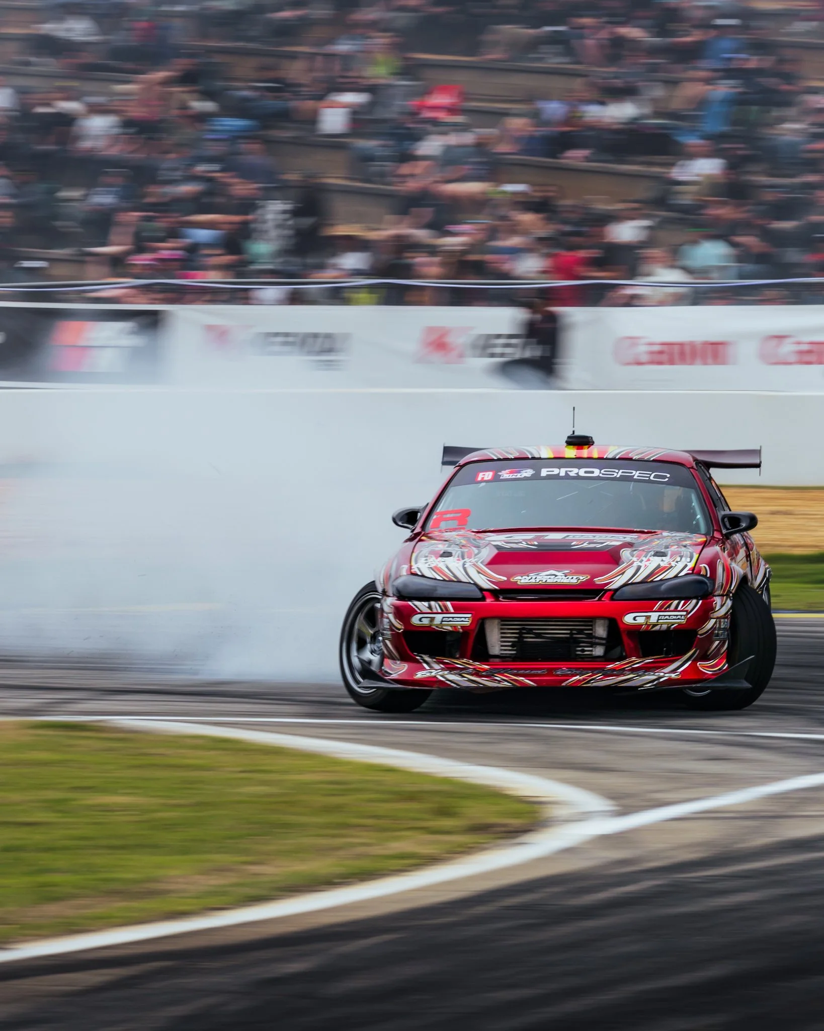 A red racing car with graphics and sponsorship decals on a race track, drifting around a corner at high speed while smoke billows from its rear tires, with a blurred crowd and barriers in the background.