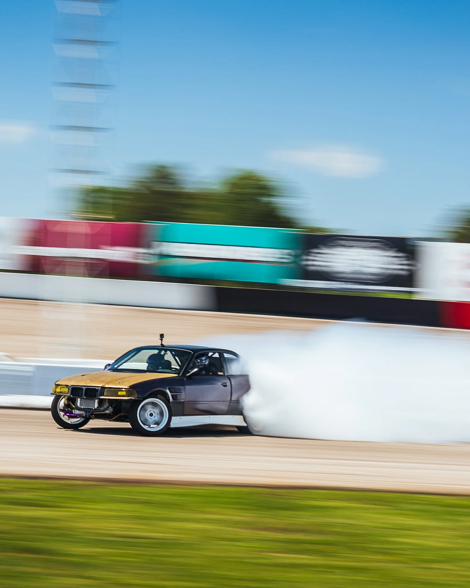 A black and yellow sports car racing on a track, with a cloud of smoke behind it, and a blurred background of trees and a racing banner.
