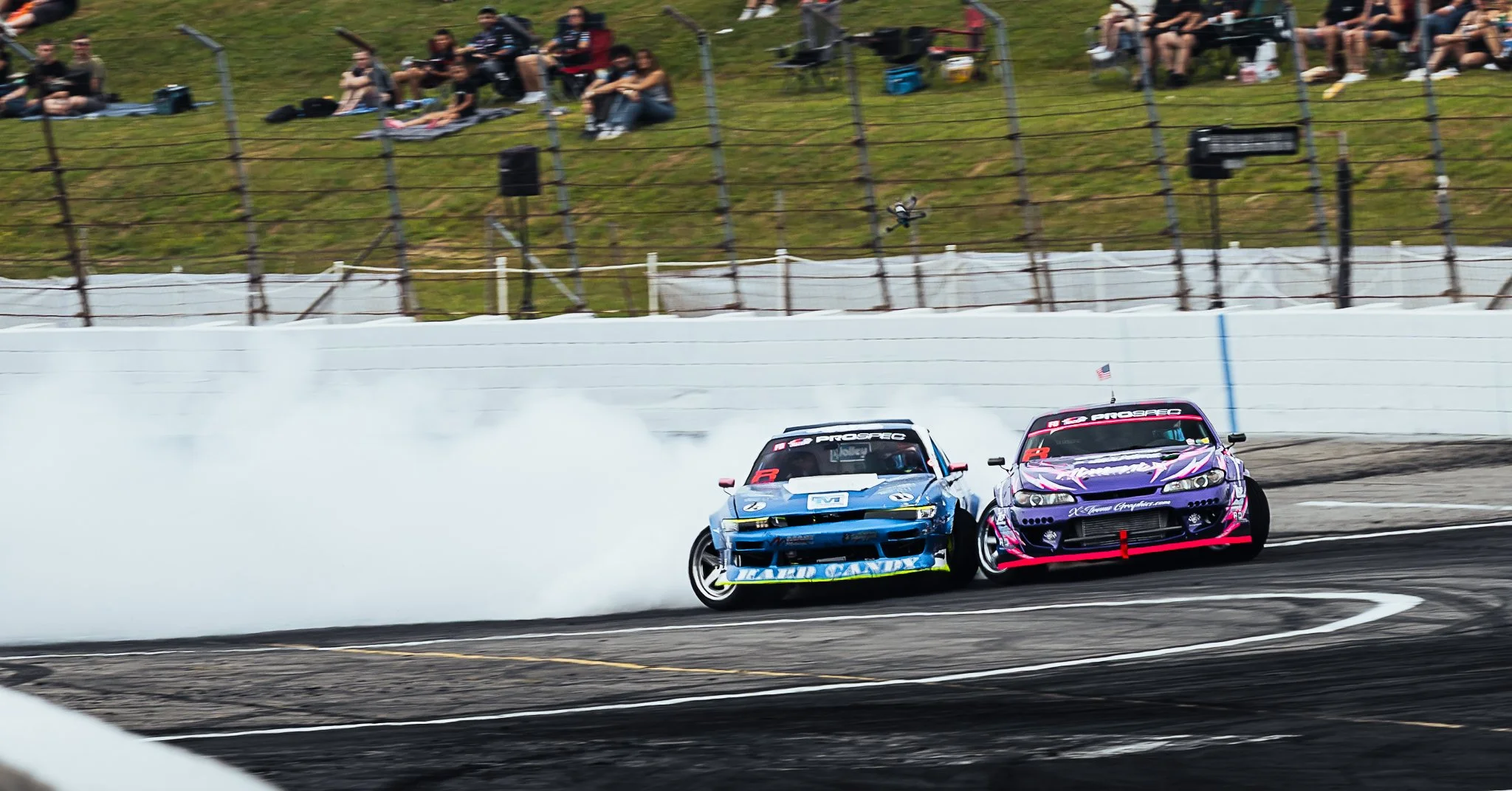 Two race cars drifting on a racetrack, creating smoke, with spectators watching from the hillside behind a fence. Formula Drift Indy