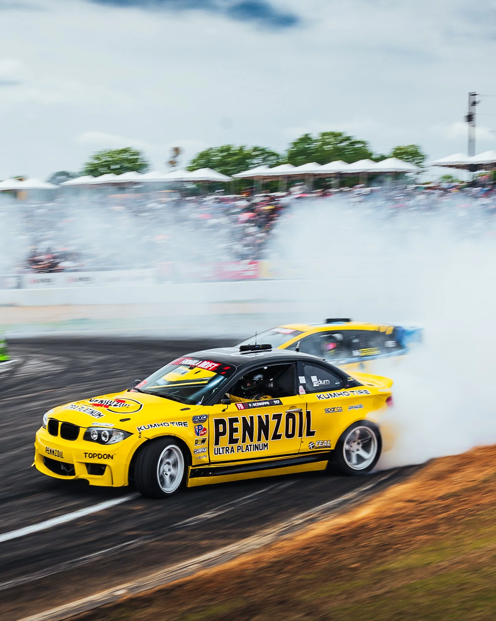 Yellow race car with Pennzoil and other sponsorship logos drifting on a race track, billowing smoke behind it.
