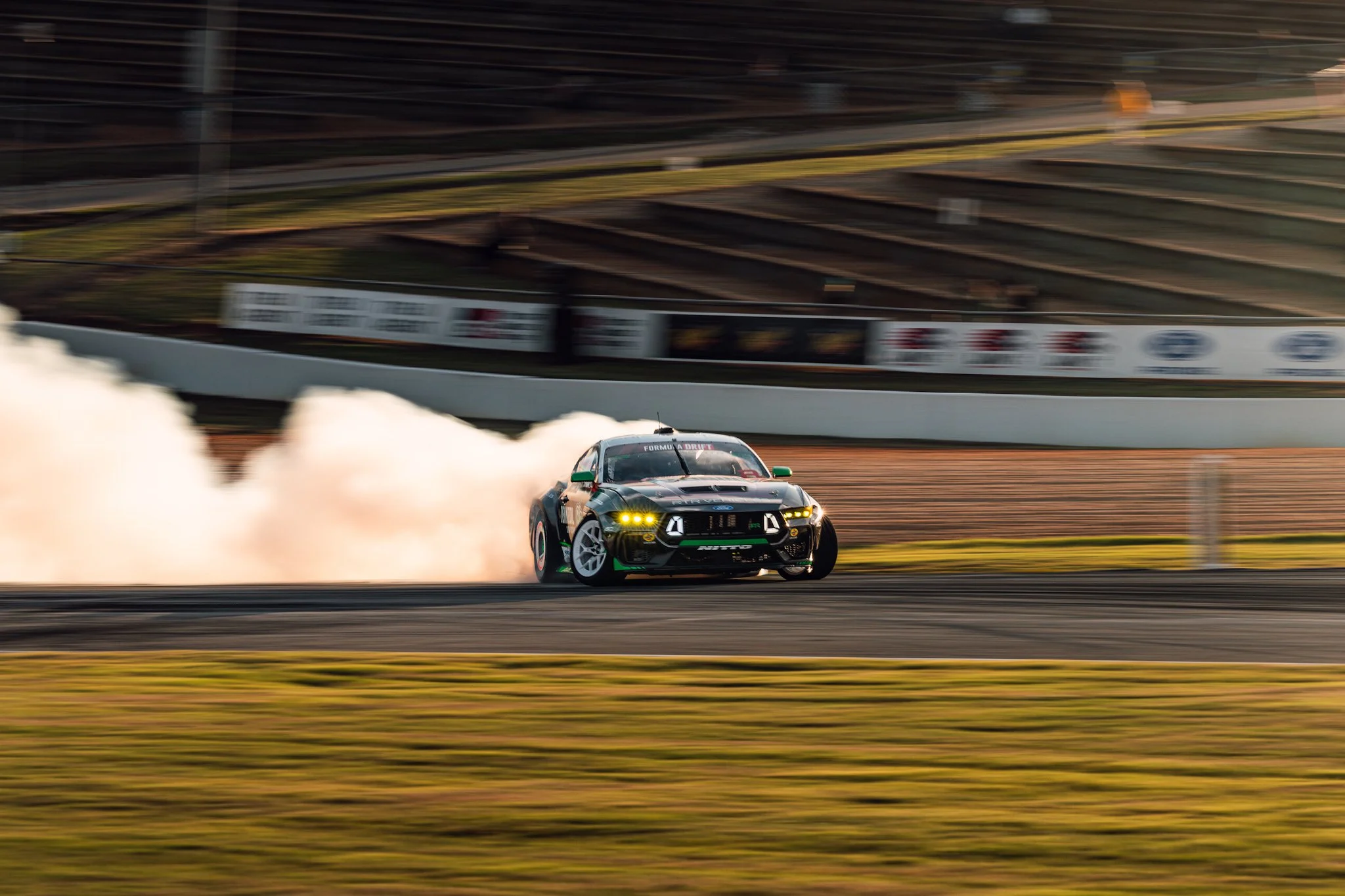 A race car drifting on a racetrack, with smoke coming from its tires and a background of empty stadium seats and advertising banners.