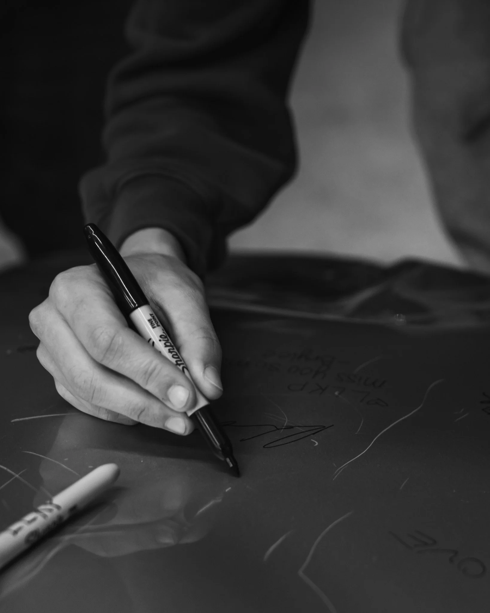 A person writing on a dark surface with a black sharpie marker, with a white marker lying nearby.