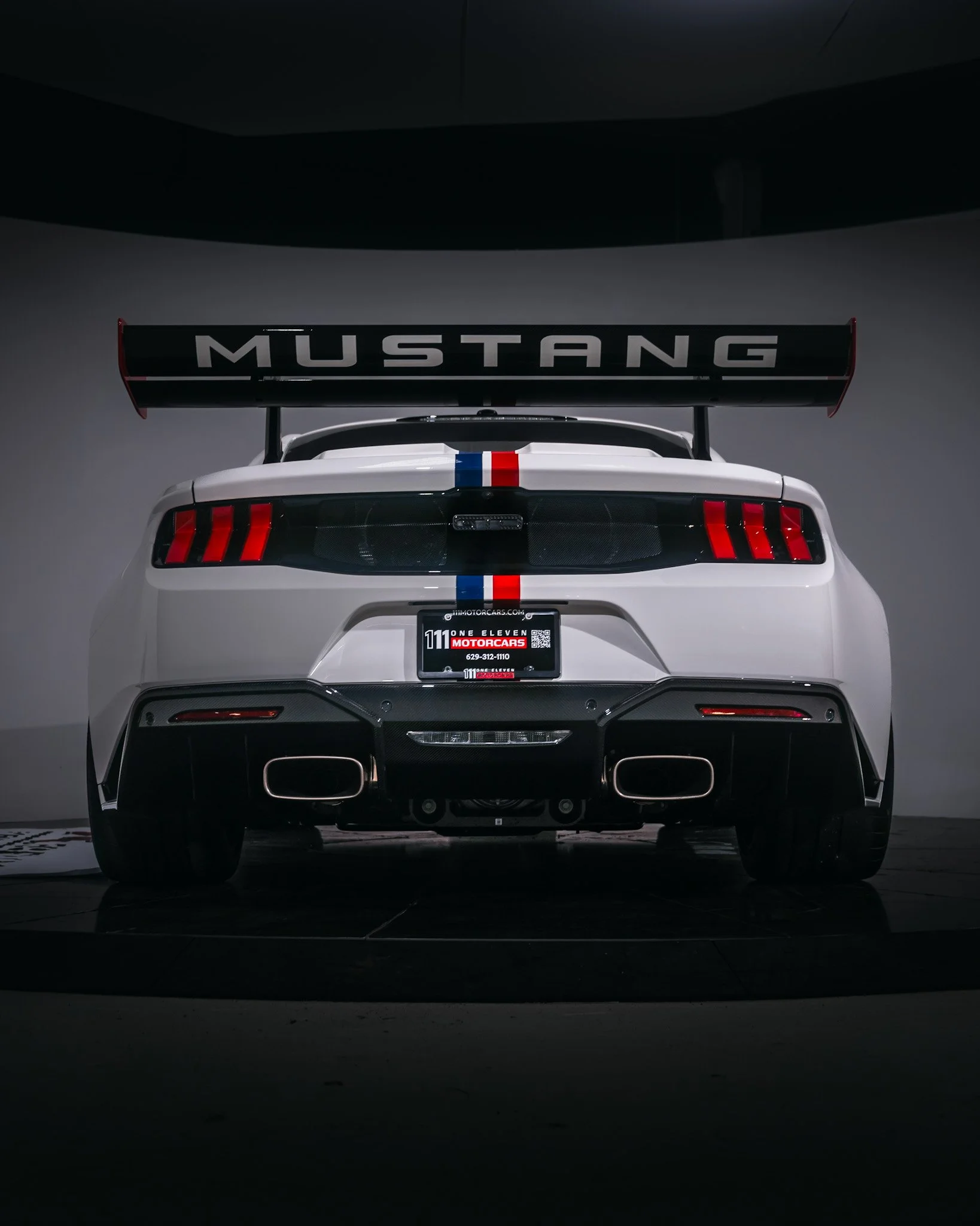 Rear view of a white Ford Mustang race car with a large rear wing and a racing stripe, in a studio setting.