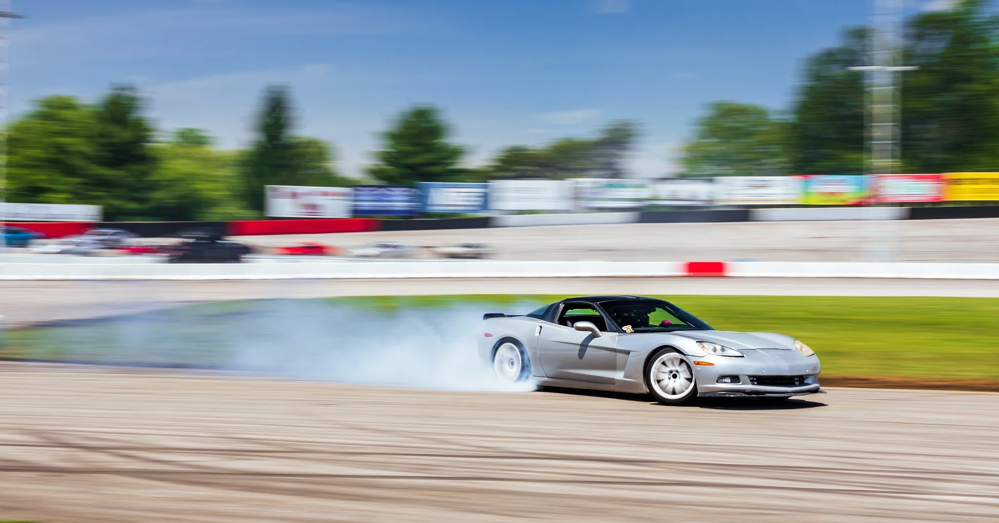 A silver sports car drifting on a race track, creating smoke from the tires, with a blurred background of trees, signage, and other cars.