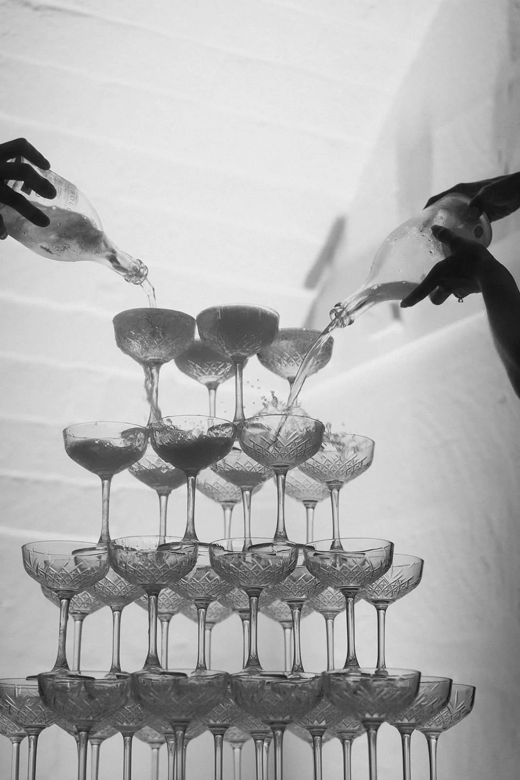 Two sets of hands pouring champagne into glasses that are stacked in a tower.