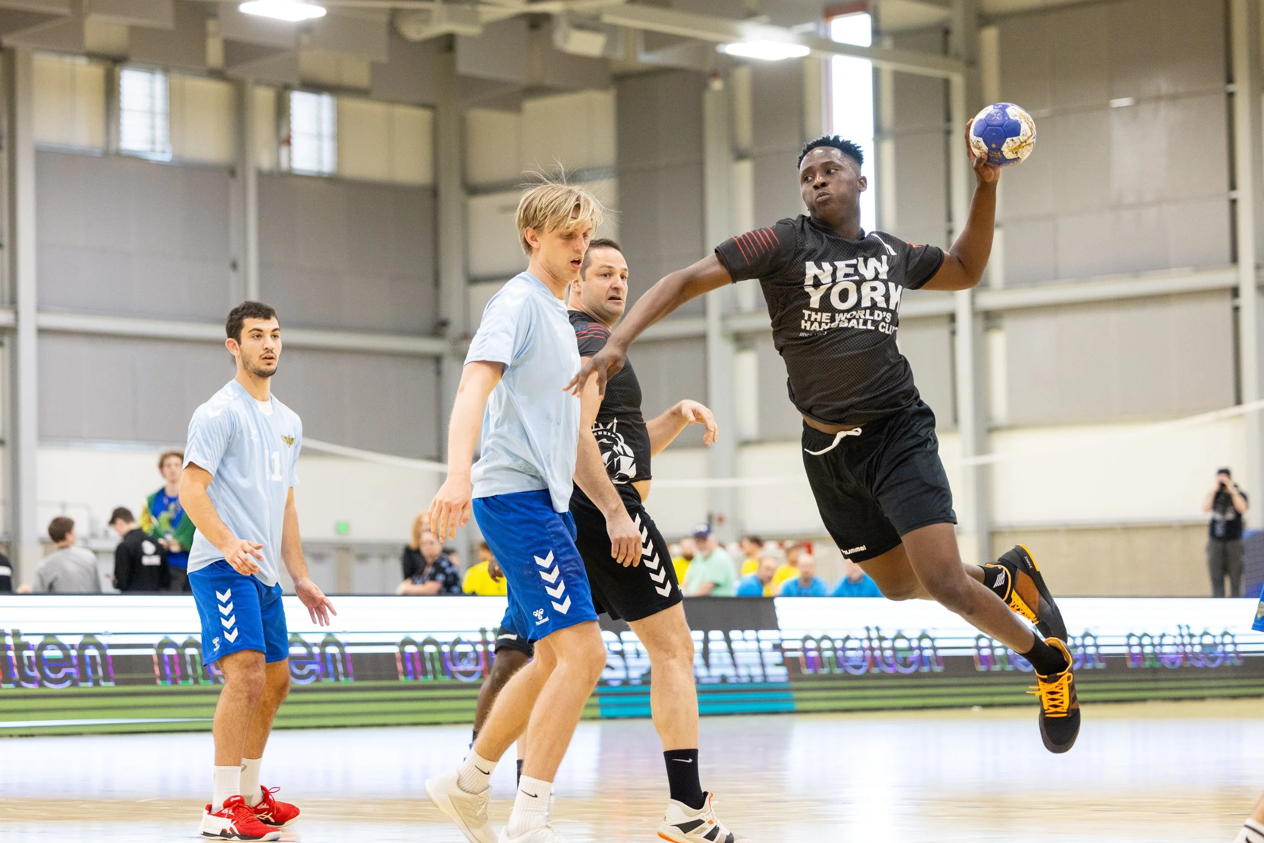 Handball game with players in motion, one player jumping while holding the ball, indoor sports arena with spectators and digital advertisement boards.
