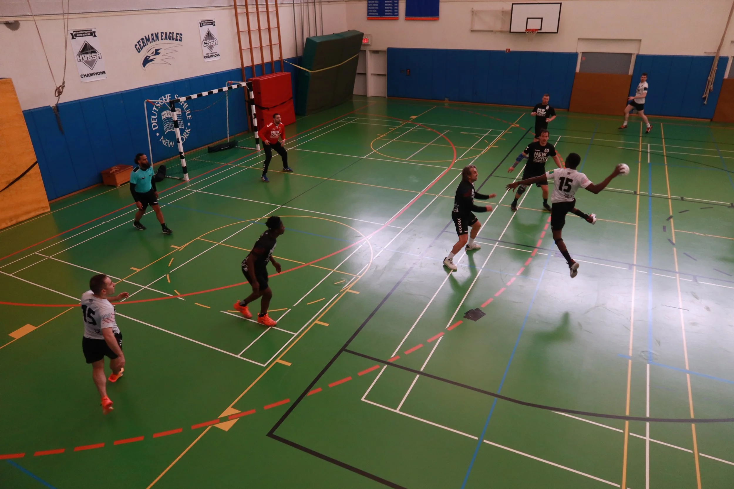 Indoor gymnasium with people playing handball, a goal in the background, and banners on the wall.