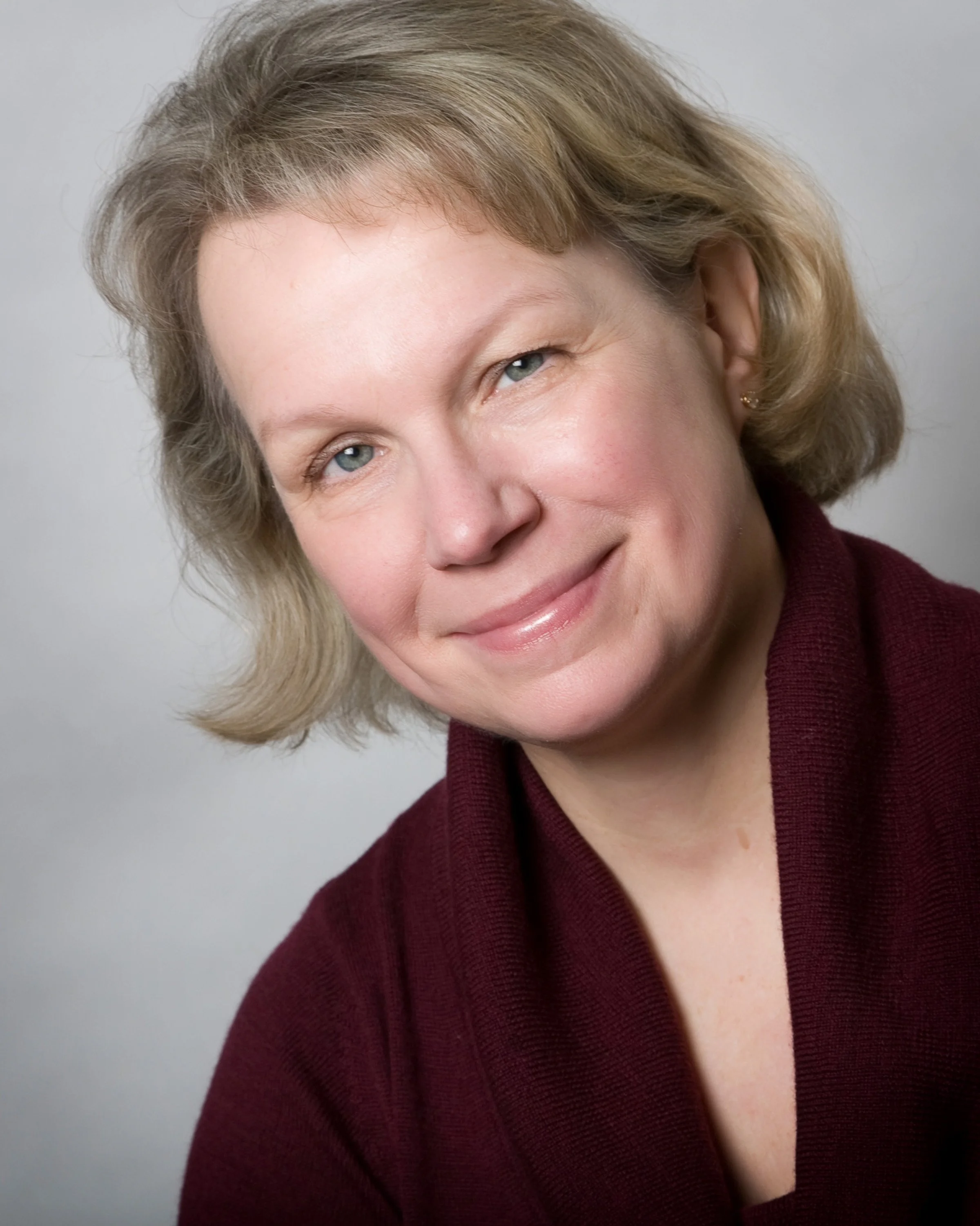 A middle-aged woman with short, blonde hair and blue eyes, smiling and wearing a burgundy top.