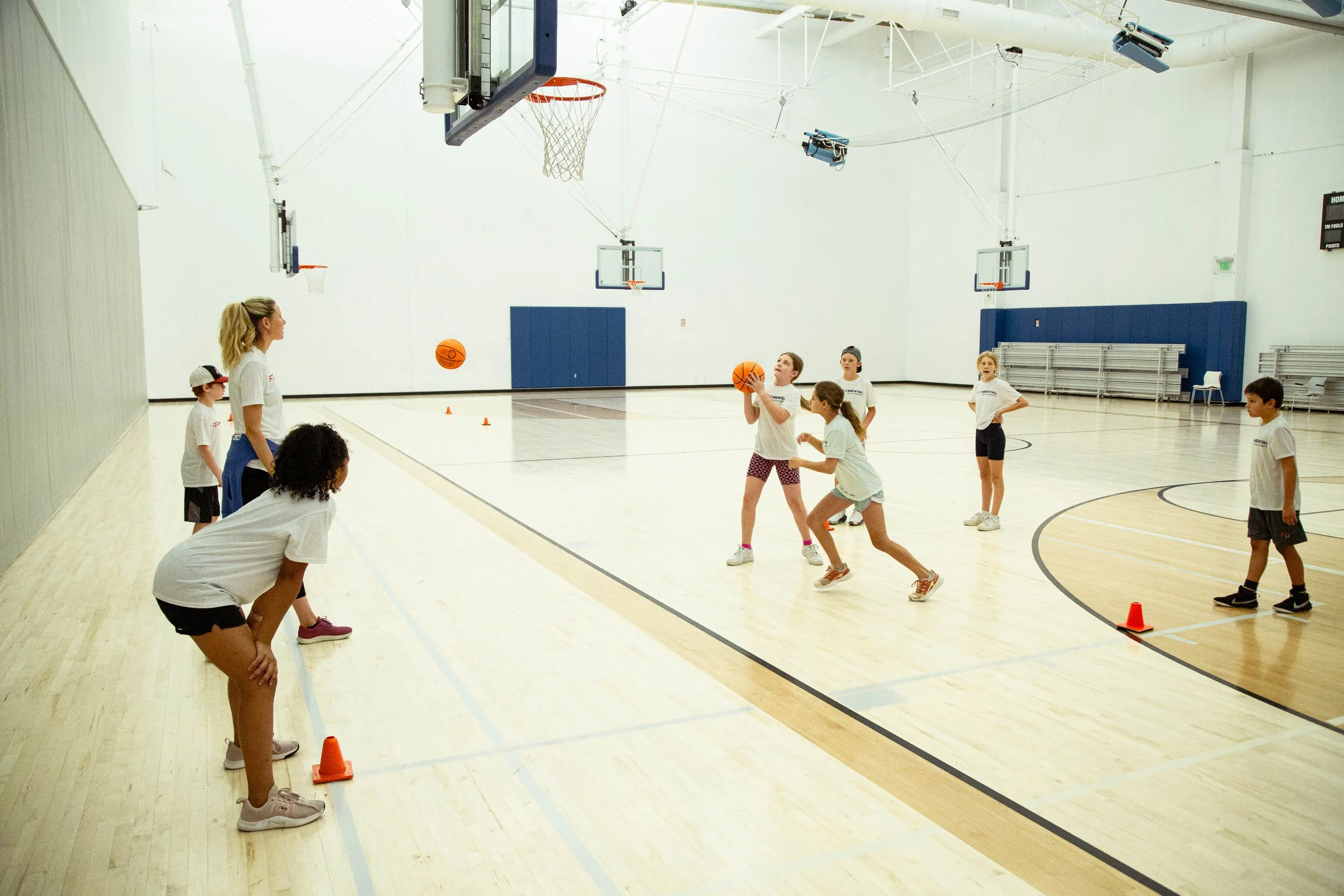 Kids and a coach playing basketball in an indoor gymnasium with a wooden floor, three basketball hoops, and a few chairs along the wall.